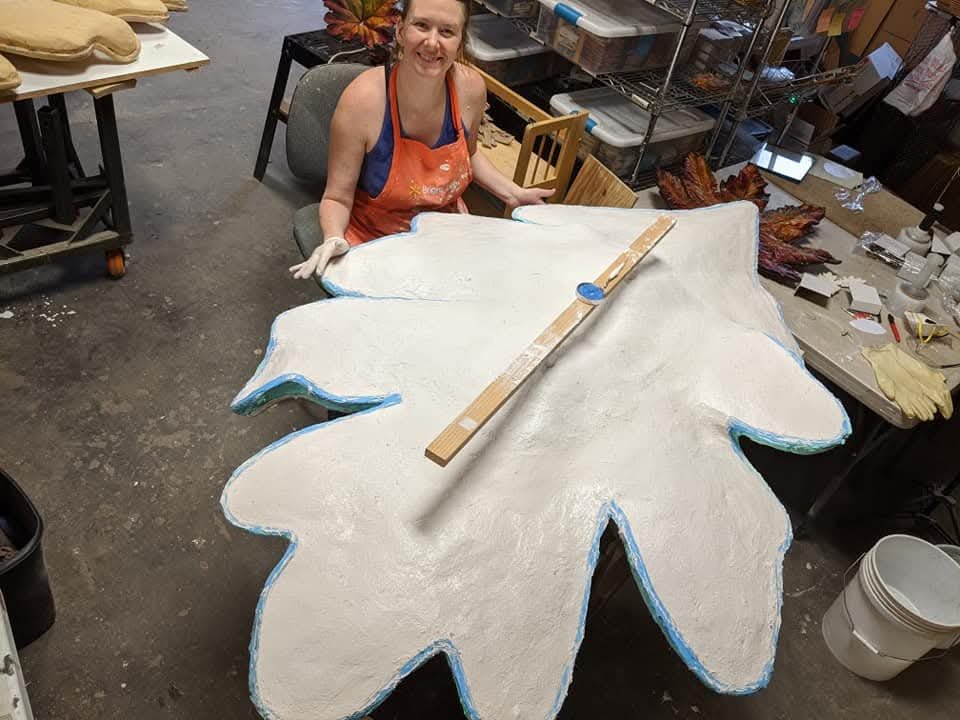 Tania McCamy wearing an orange apron and gloves is sitting at a table in a workshop, smiling at the camera. In front of her is a large, flat, leaf-shaped object  with a ruler placed on it. The art studio can be seen in the background.  