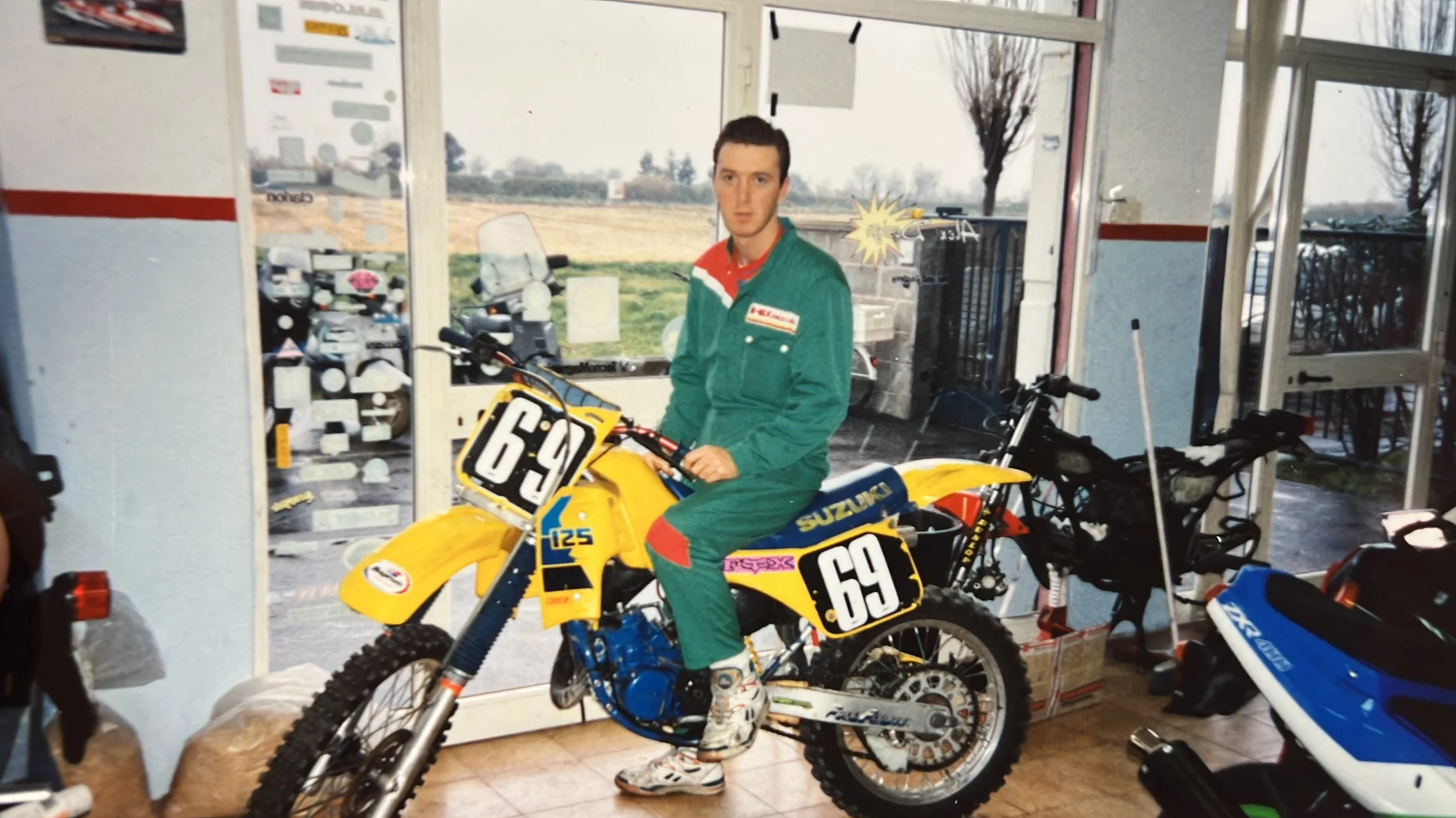 Un giovane Gianni in tuta da motocross seduto su una moto da corsa gialla con il numero 69, all'interno di un’officina o negozio di moto.