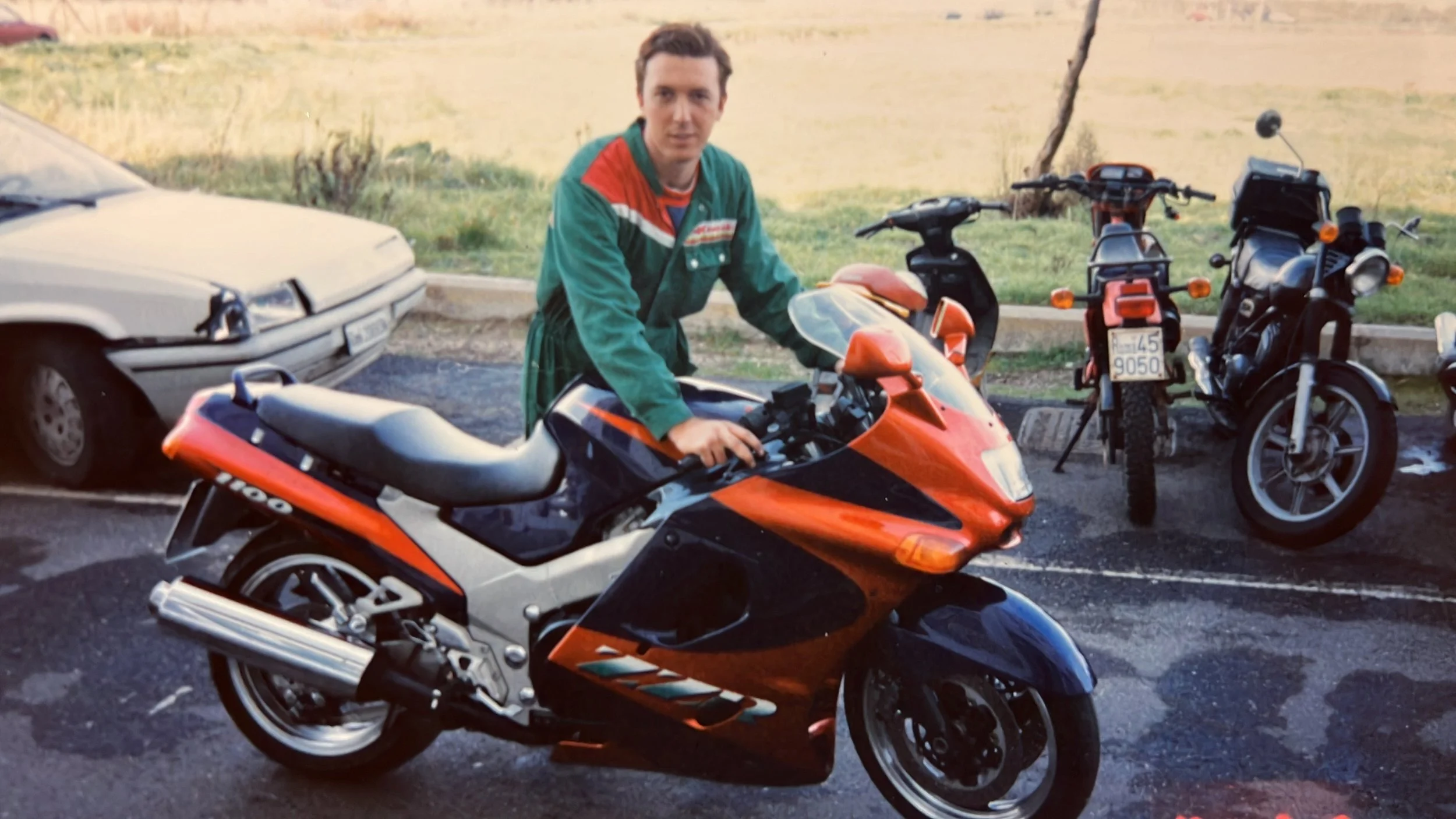 Un uomo con giacca verde si trova vicino a una motocicletta sportiva arancione e nera, con altre due moto alle sue spalle, e un'auto bianca parcheggiata vicino.
