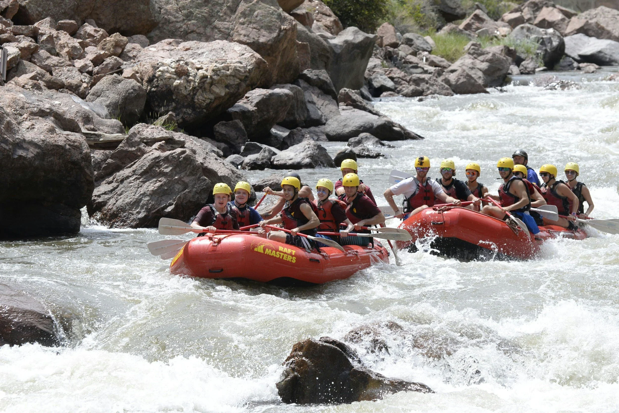 Group of people white water rafting on a river with rocks and trees in the background, all wearing helmets and life jackets.