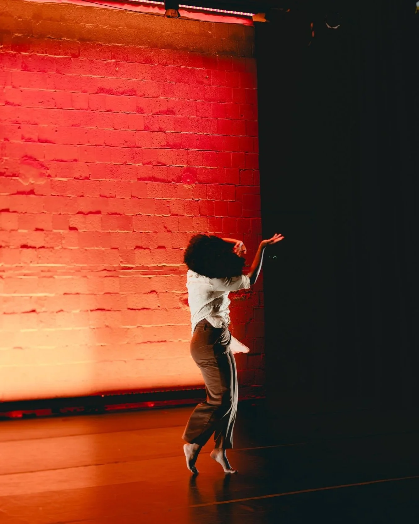 A woman dancing on a stage with a red brick wall illuminated by red and orange lighting, wearing a white top and beige pants.