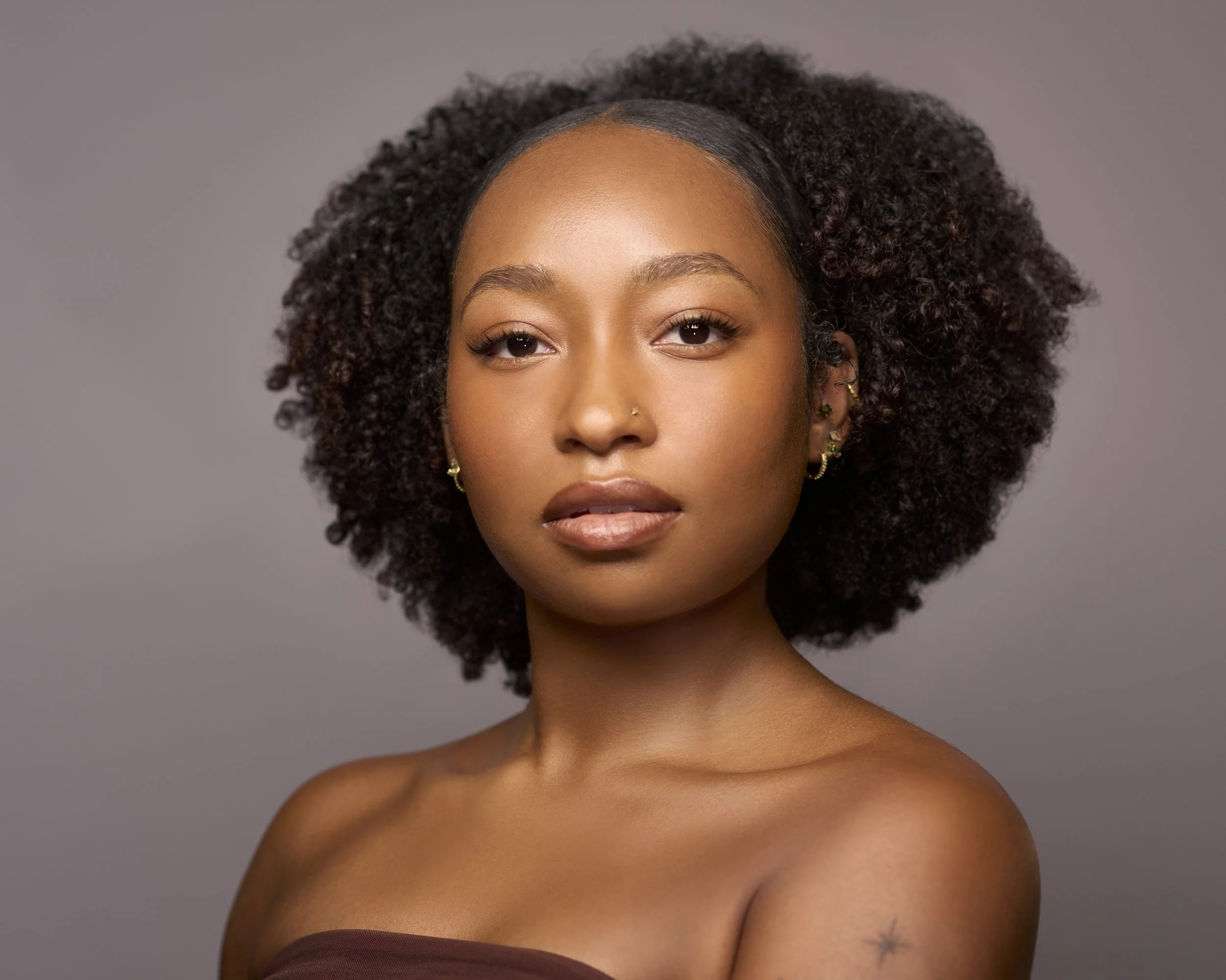 Close-up portrait of a woman with natural curly hair, wearing earrings, with a neutral background.
