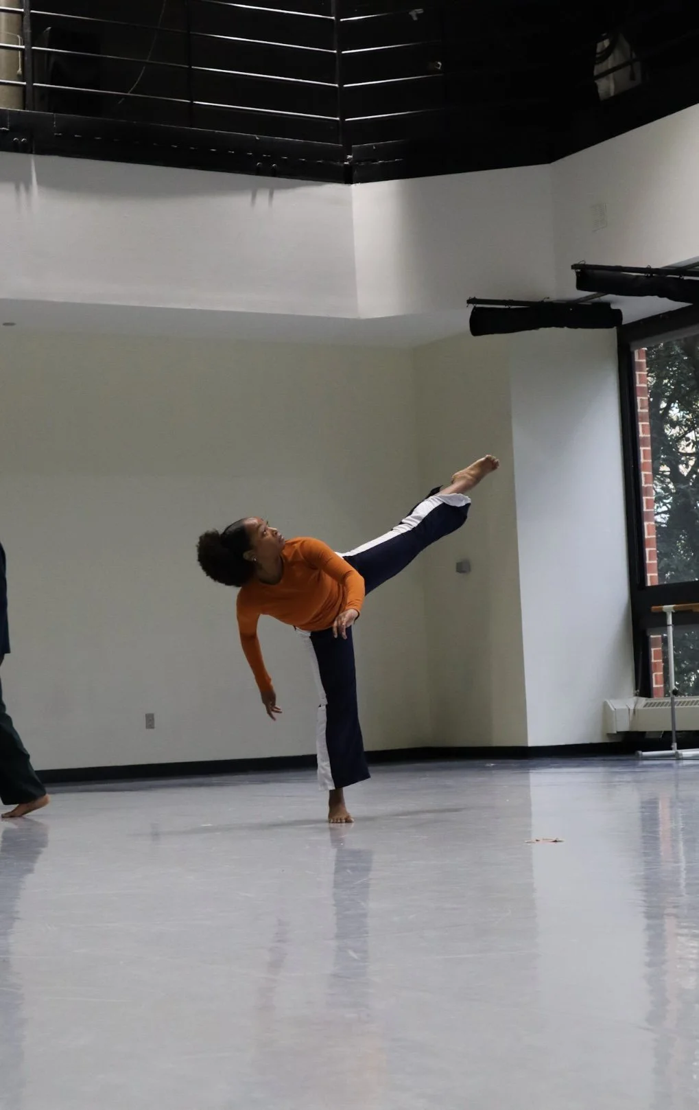 A woman practicing a high kick in a spacious room with large windows and a ballet barre.