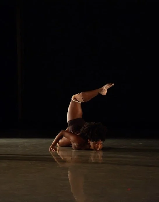 A female dancer performing a pose on stage with a black background, one leg raised and bent over her head.