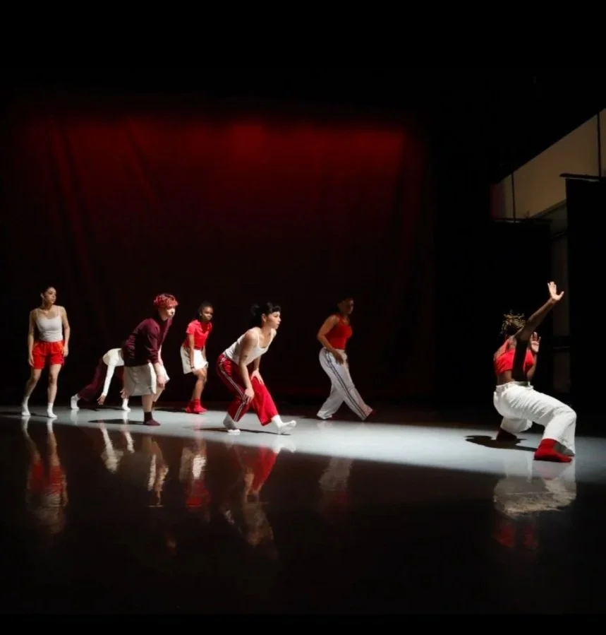 A group of seven dancers on stage, practicing or performing a routine, with one dancer kneeling and raising her arm, dressed in rehearsal clothes.
