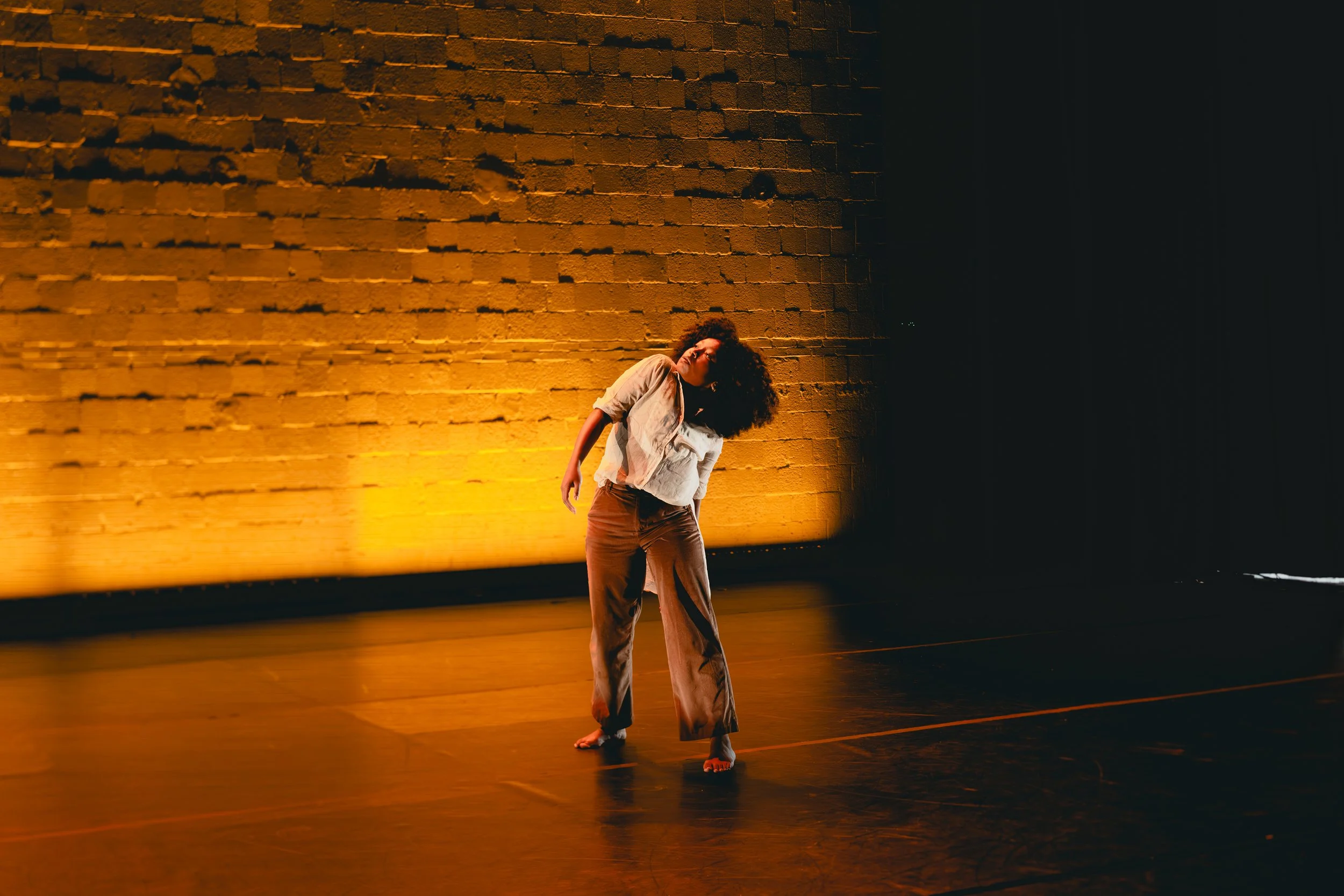 A woman with curly hair and a light-colored blouse and pants is dancing barefoot on a dimly lit stage with a large brick wall background illuminated in warm yellow-orange light.