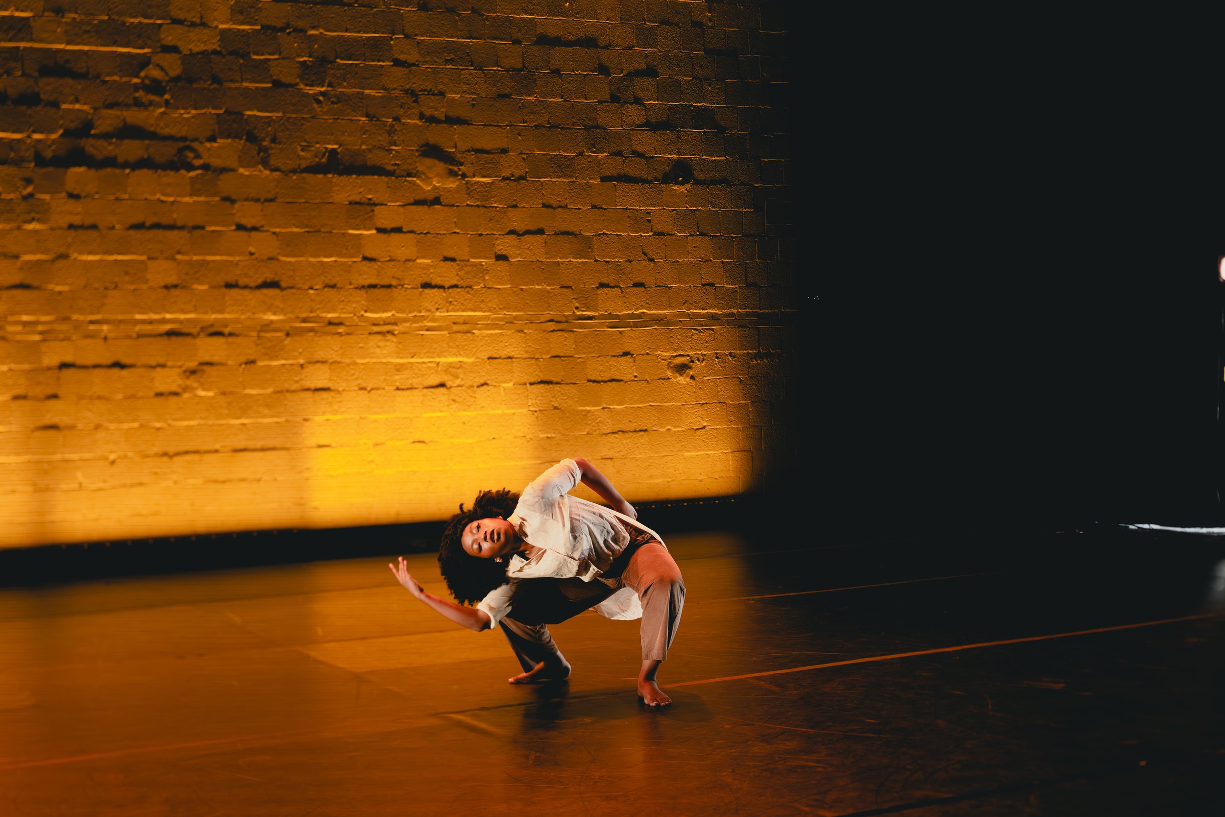 A dancer with curly hair performing on stage against a brick wall illuminated by warm yellow lighting. The dancer is in a low, stretched pose with one arm reaching upward and the other arm bent near her face.