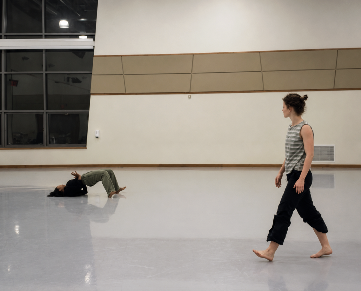 Two women practicing dance or yoga in a spacious, well-lit studio with minimal decor. One woman is lying on her back with her knees bent, and the other is walking barefoot.