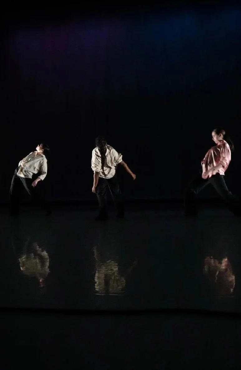 Three dancers on dark stage with reflective floor, minimal lighting, and a dark background.
