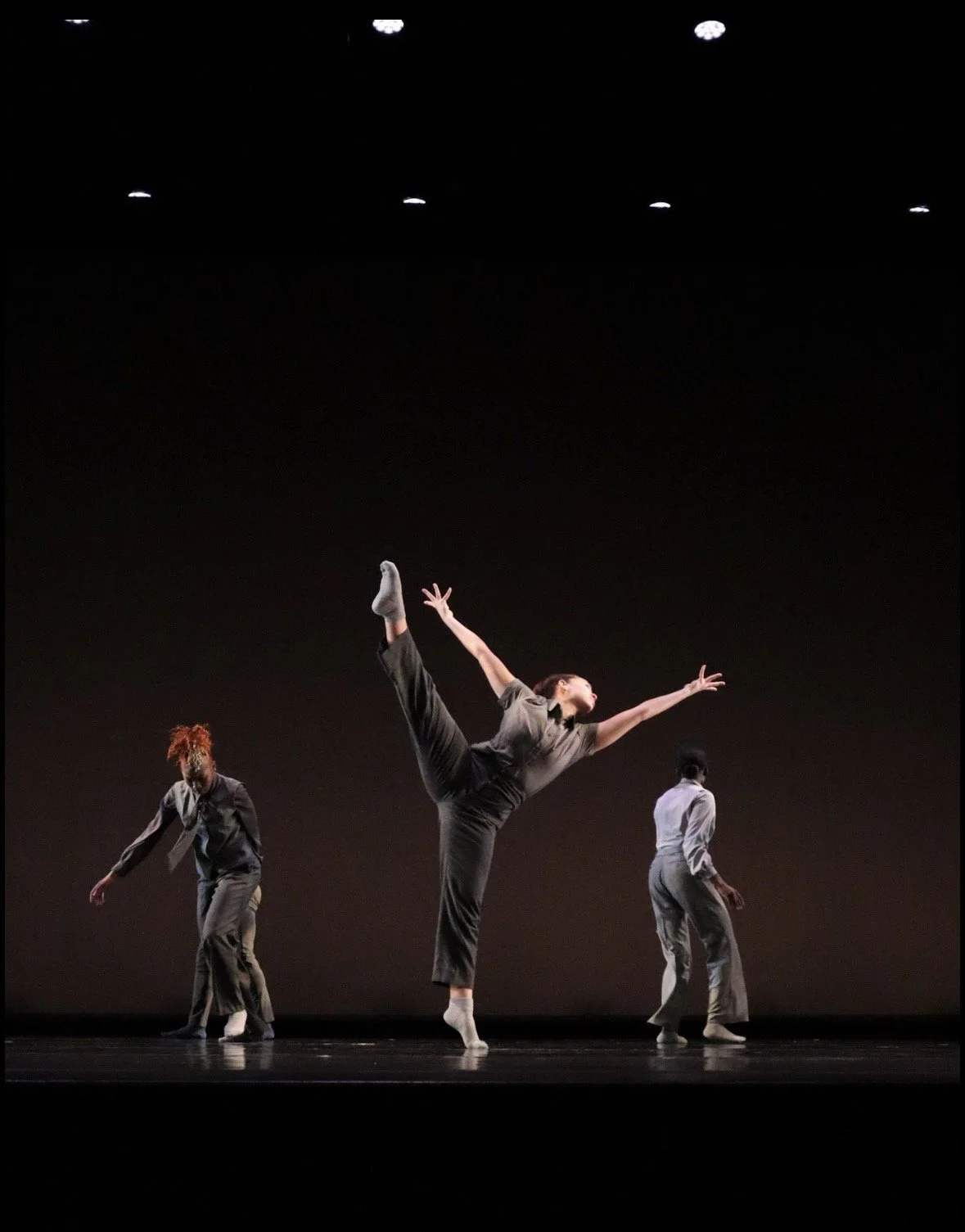 Three dancers perform on a dark stage, with one dancer in the center executing a high kick while the other two dancers move in different directions.