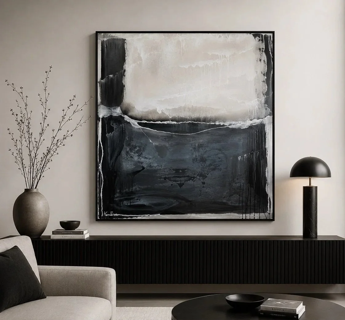 Modern living room with a large black and white abstract painting on the wall, a black sideboard with a black table lamp and a large gray vase with branches, a small bowl, and books, and a round black coffee table with a black bowl and books.