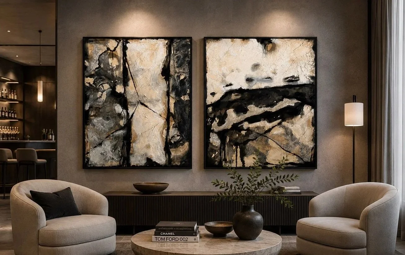 Modern living room with abstract black, white, and beige paintings, two beige armchairs, and a round coffee table with books and vases.