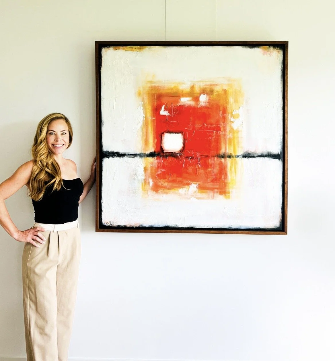 A woman with long blonde hair wearing a black top and beige pants standing next to an abstract painting with red, orange, yellow, black, and white colors on a white wall.