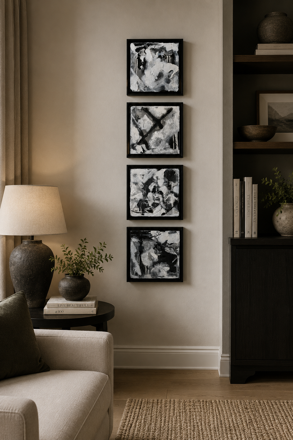 A living room corner with a beige sofa, a dark side table with two books and two plant pots, a table lamp, and a black and white abstract art piece of four panels hanging on the wall. There is a bookshelf with decorative items and a plant to the right.