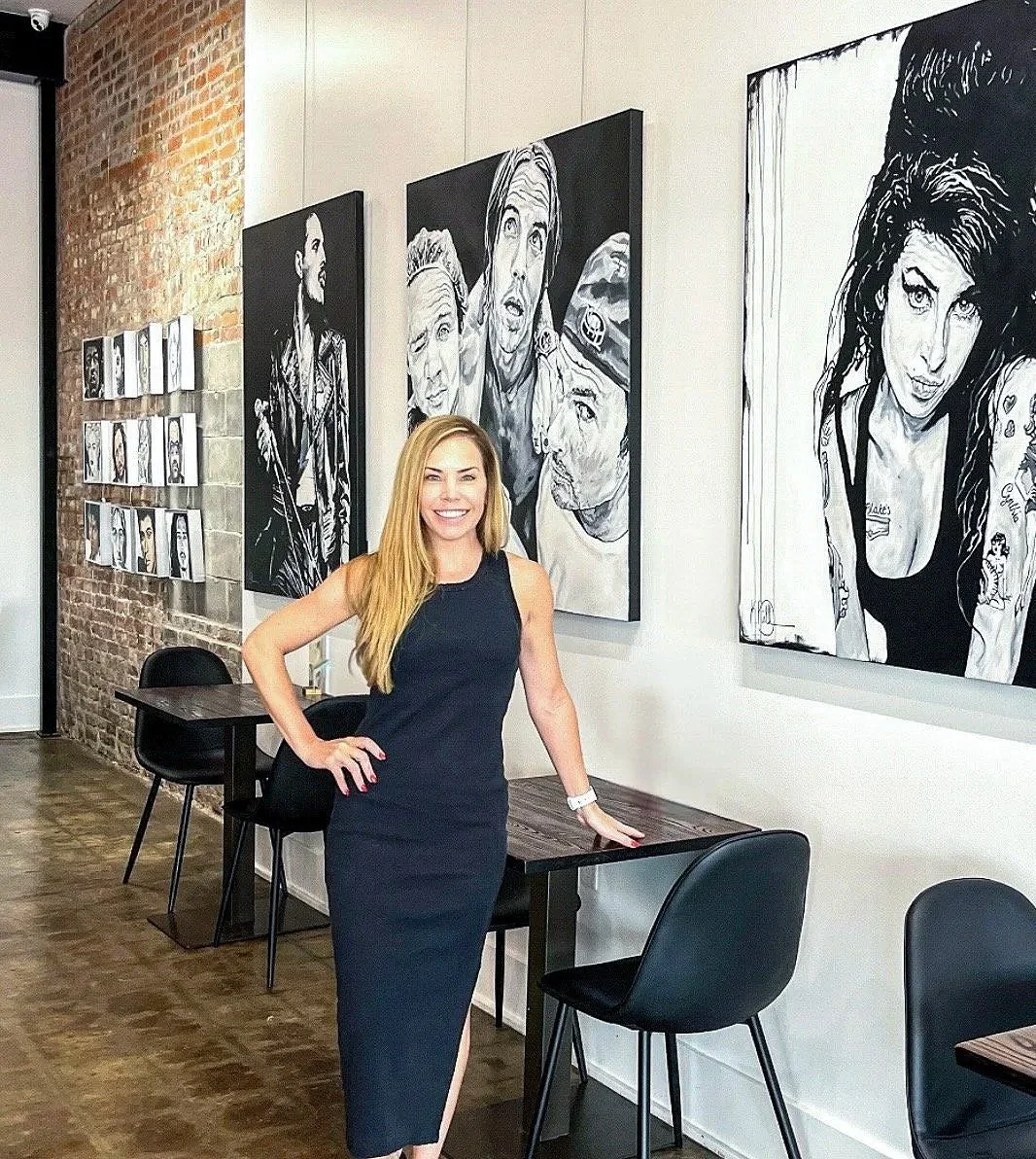 A woman with long blonde hair in a sleeveless black dress stands inside an art gallery or cafe, smiling at the camera. Behind her, there are black and white portraits of various people hanging on the white wall and a brick wall with smaller artworks.