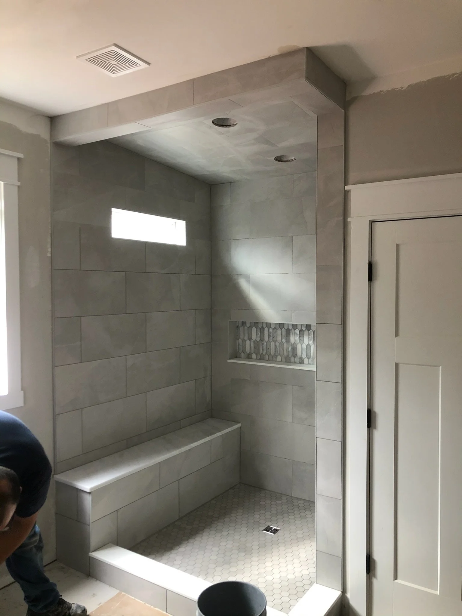 A partially constructed or renovated walk-in shower with large rectangular gray tiles, installed bench, niche with hexagonal patterned tiles, and small hexagonal floor tiles. There is also a small rectangular window, ceiling recessed lights, and a floor drain.