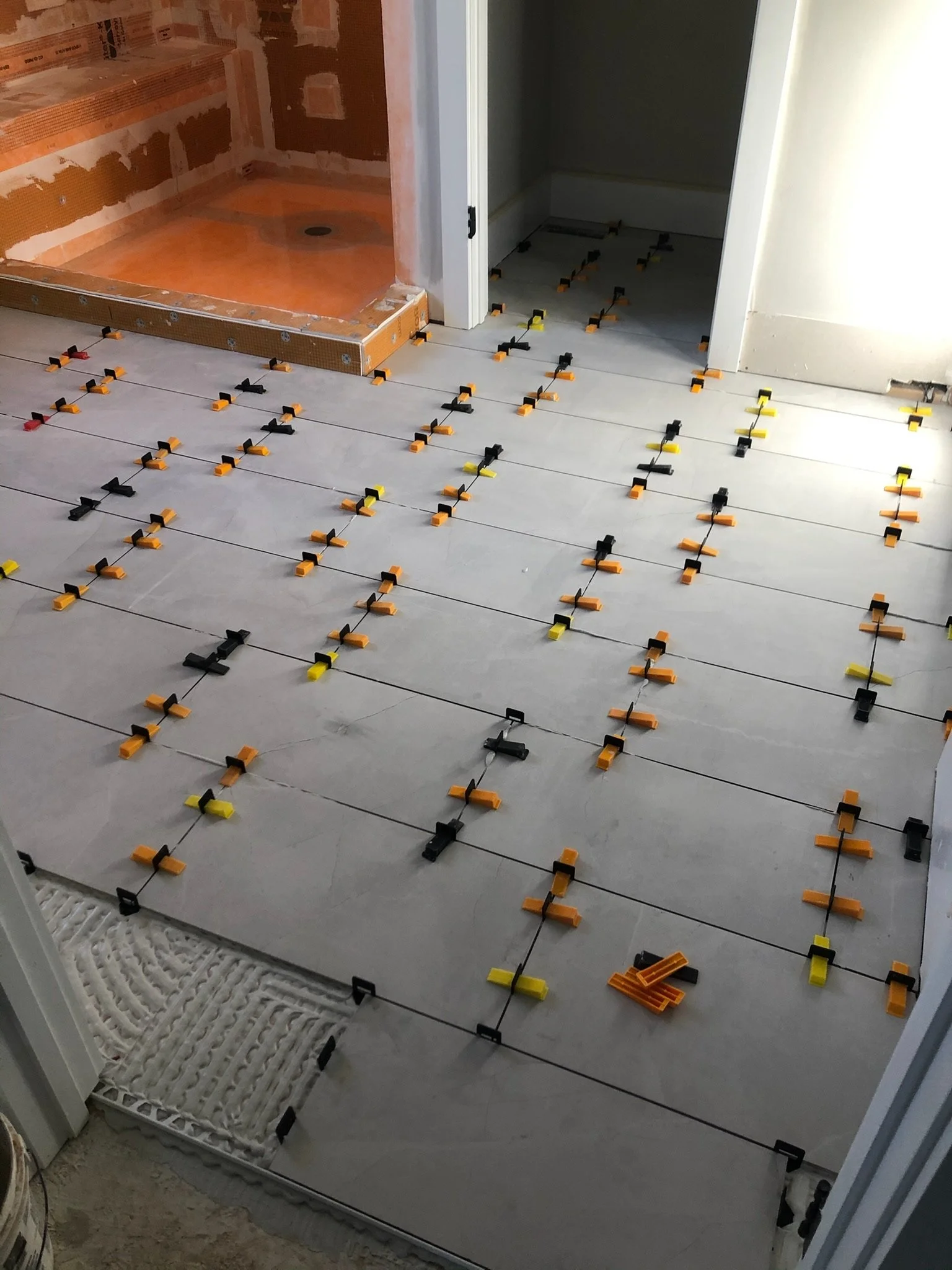 Floor installation process with spacers and tiles in a bathroom under construction, showing a door leading to a shower area with visible plumbing.