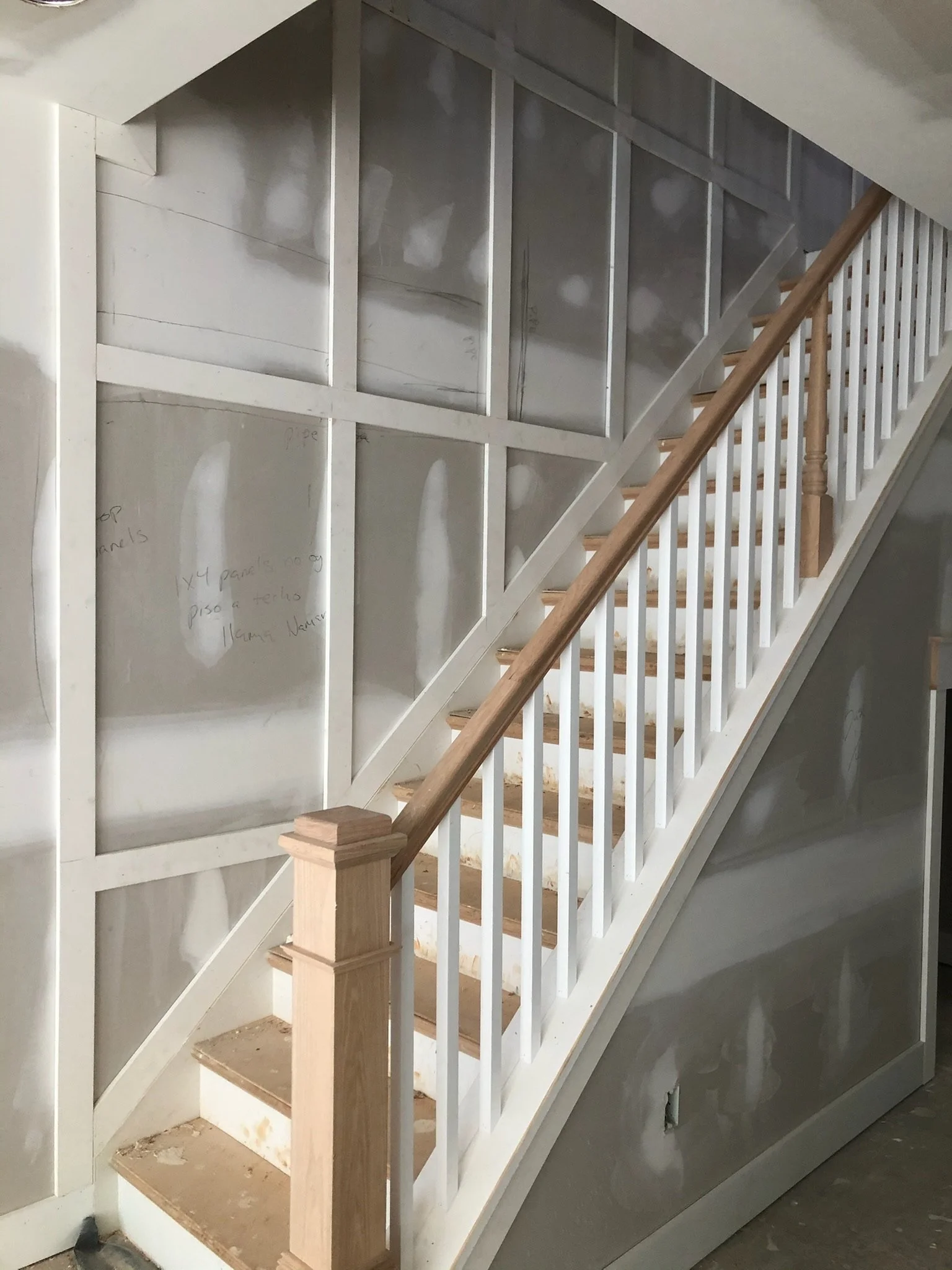 Interior view of a staircase with unfinished details, showing a wooden handrail and white balusters, alongside a partially completed wall with notes written on it.