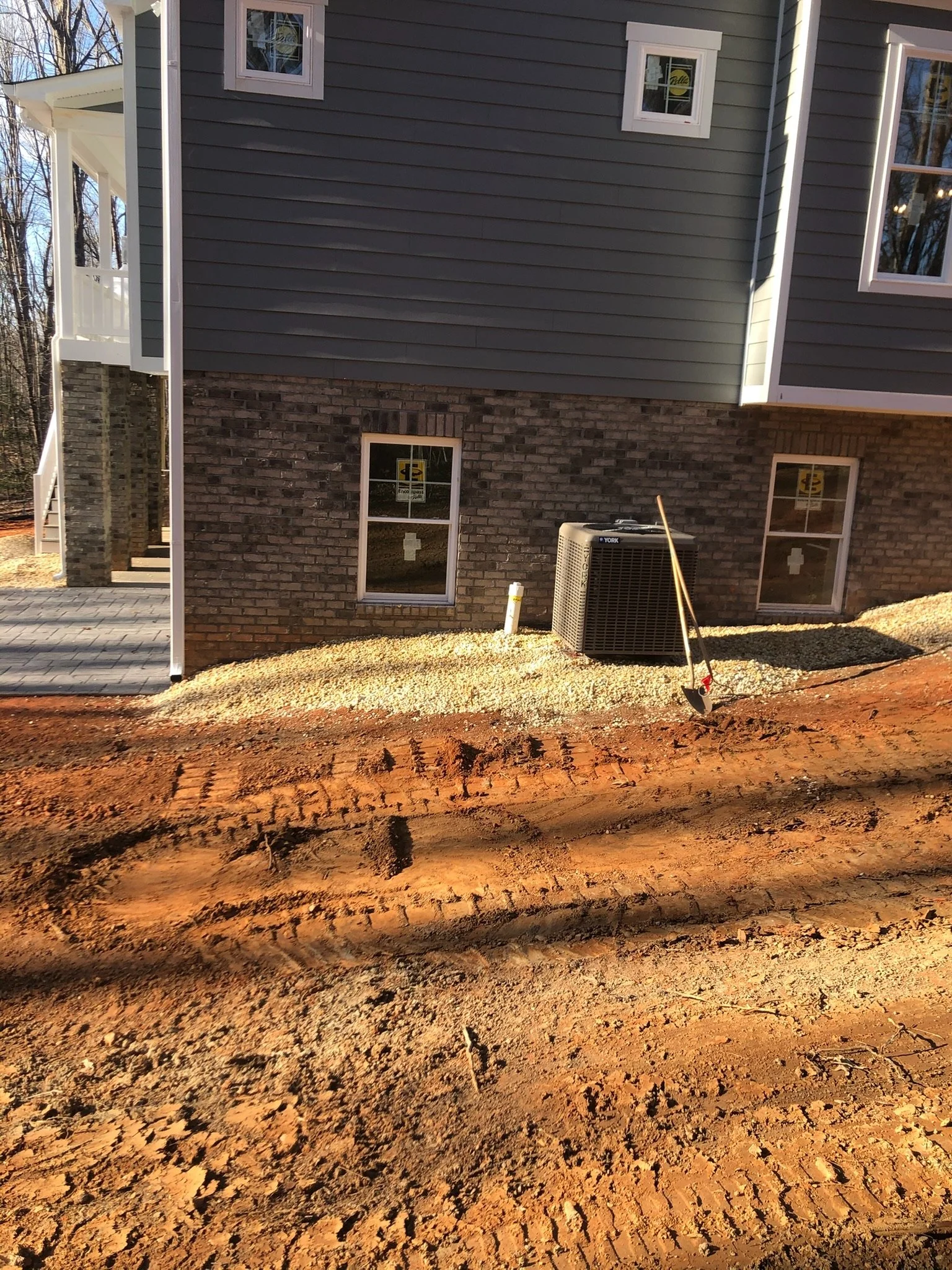 Photograph of the exterior of a house, showing brick and siding, with windows, an HVAC unit, and a shovel in the soil in the foreground. The ground appears to be under construction or landscaping.