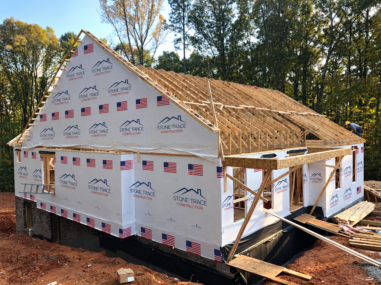 A house under construction with its wooden framework and roof support in place, surrounded by trees and construction materials.