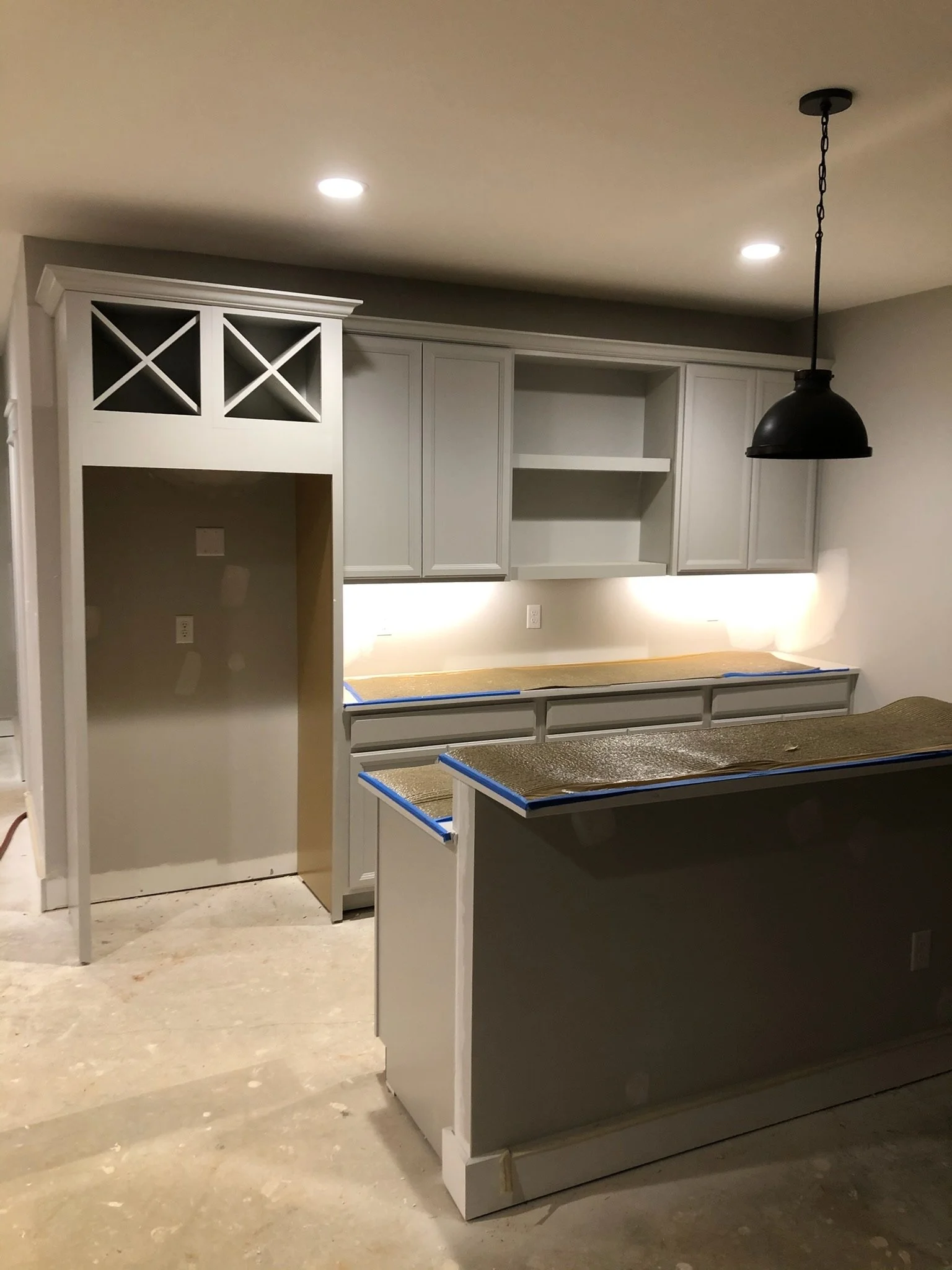 Unfinished kitchen with gray cabinets, missing countertops, and an island, with a black pendant light hanging from the ceiling and two recessed lights in the ceiling.