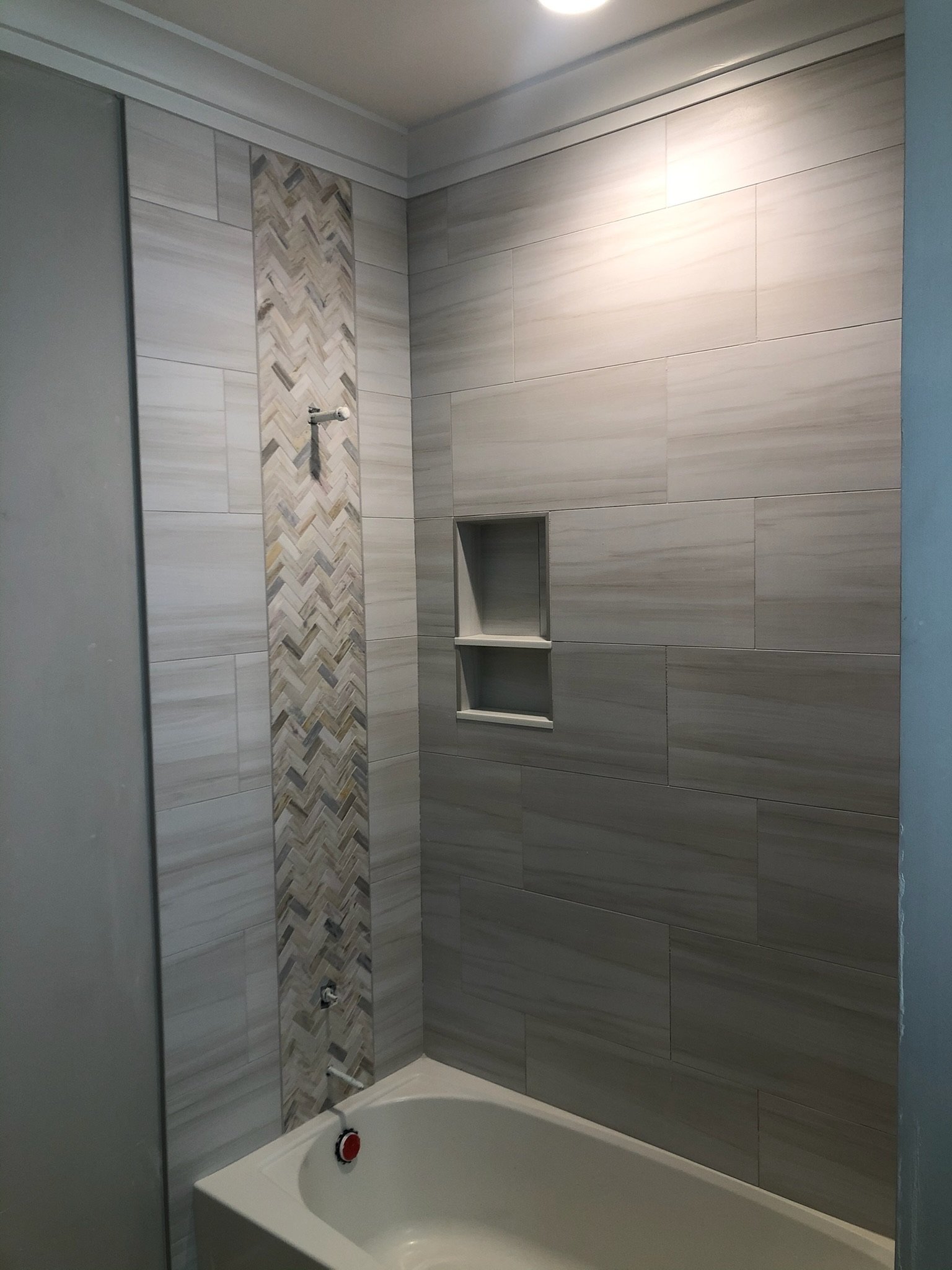 This is an image of a bathtub in a bathroom with tiled walls. The wall tiles have a modern, neutral color scheme with a decorative vertical strip of chevron-patterned tiles. There is a small built-in shelf in the wall for toiletries, and a showerhead