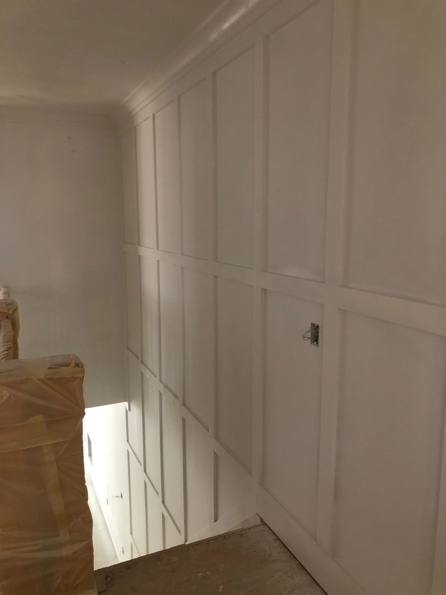 A newly installed white paneled cabinet wall in a room under construction, with a small opening for electrical wiring and a cardboard box on the floor.