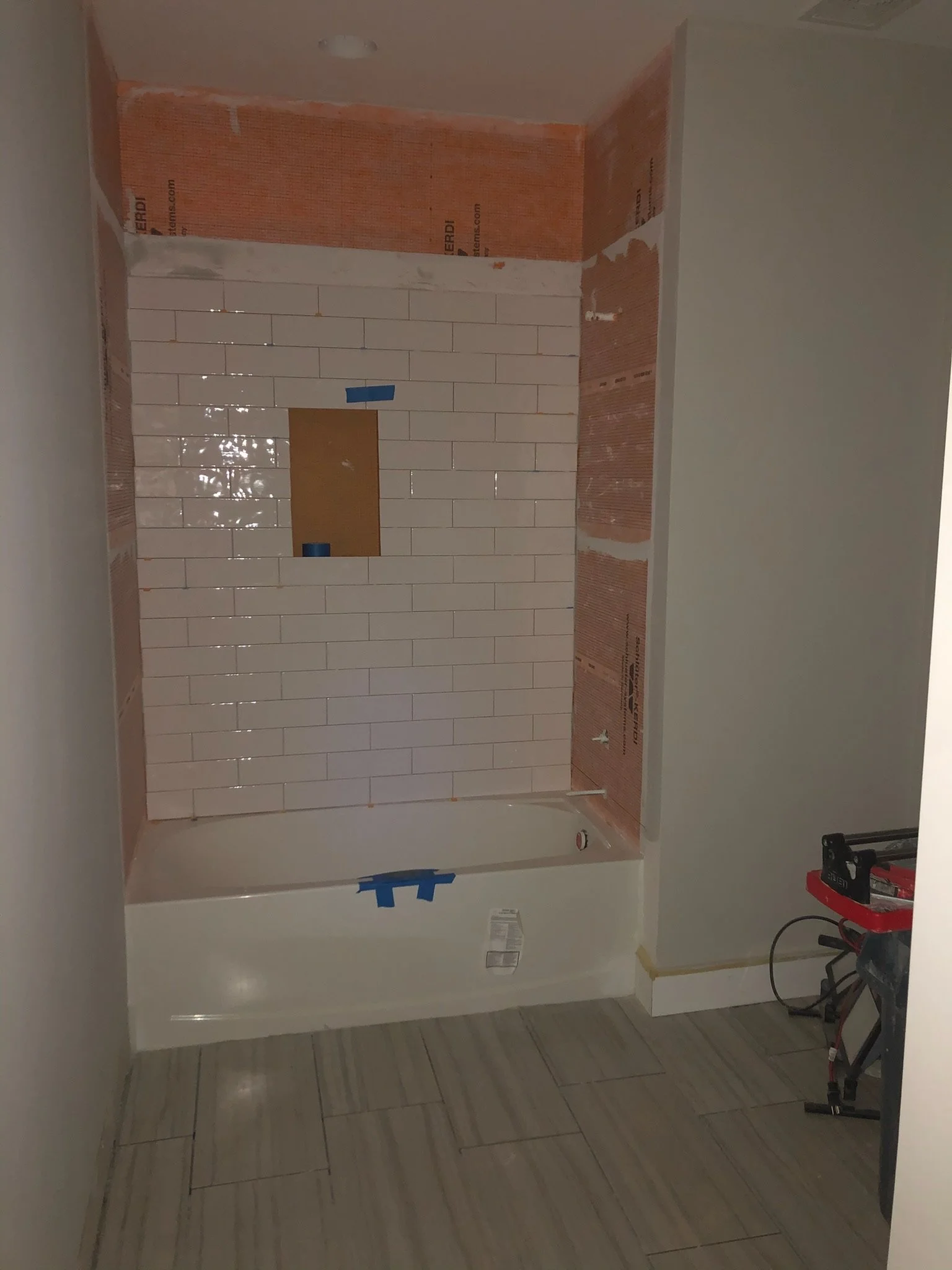 Bathroom under renovation with partially tiled wall, exposed drywall, and construction tools on the floor.
