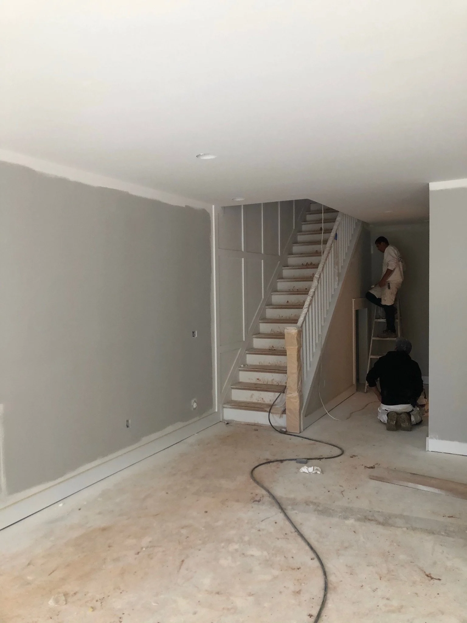 Interior of a house under renovation with a staircase, a man working on the wall, and another person kneeling on the floor, surrounded by construction tools and materials.