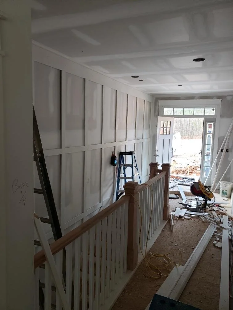 Interior of a house under construction with unfinished drywall, a blue ladder, construction tools, and debris on the floor near an open front door.
