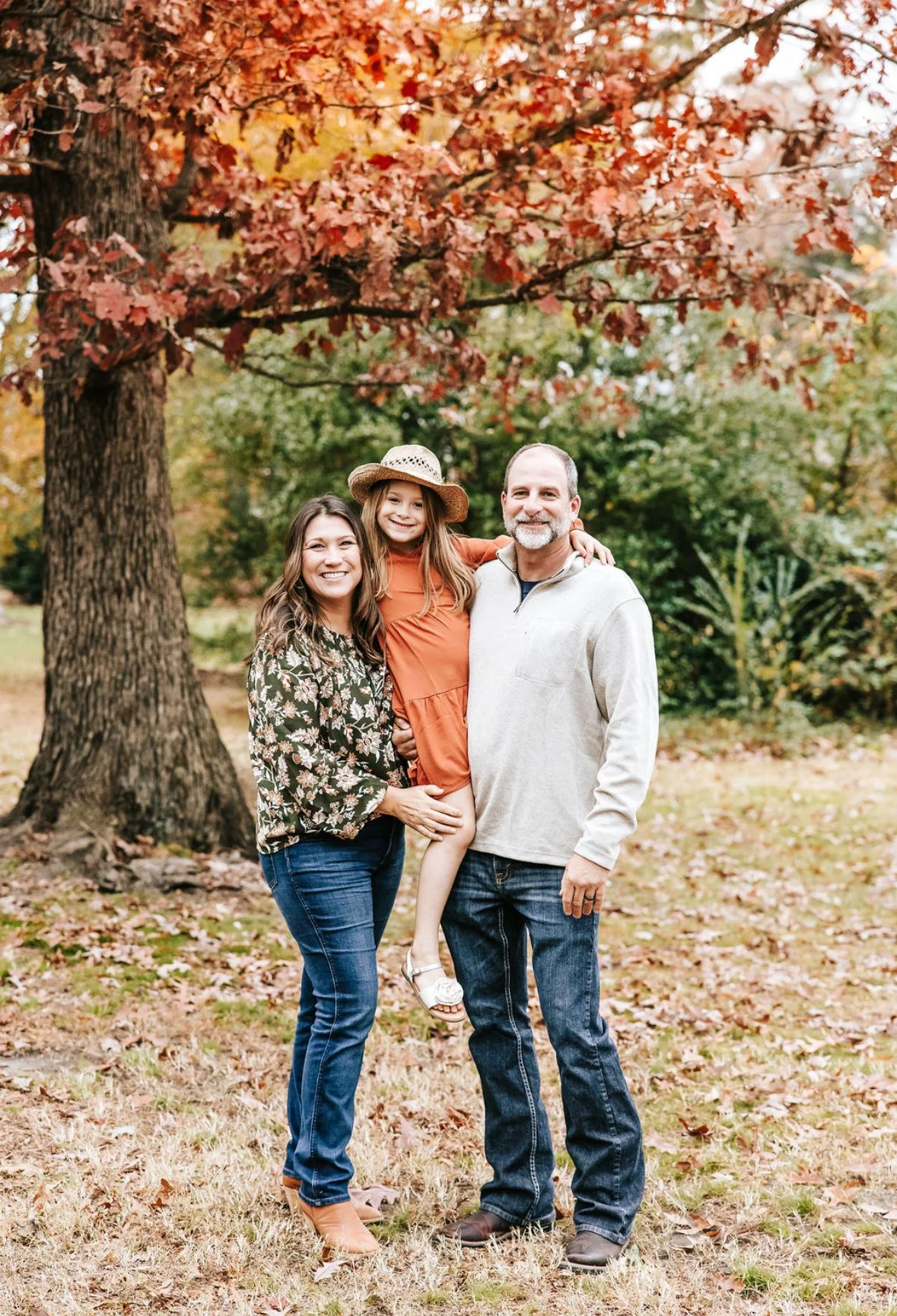 A family of three standing outdoors near a large tree with autumn-colored leaves, on a grassy area with fallen leaves. The woman is holding a young girl in an orange dress and straw hat, and the man stands beside them, all smiling at the camera.