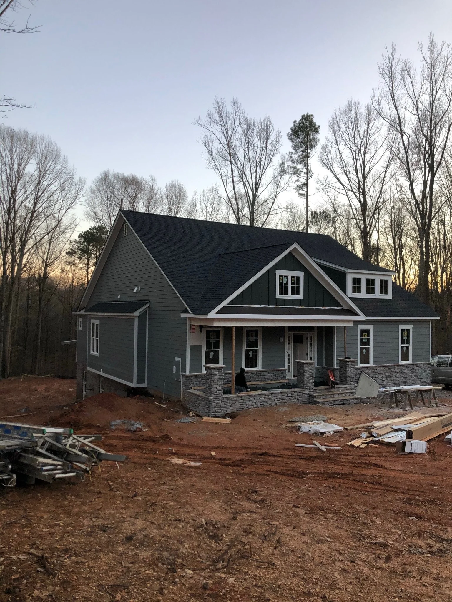 A house under construction with a grey exterior, black roof, multiple windows, and a stone foundation, surrounded by construction materials and dirt ground, with trees and a clear sky background at sunset.