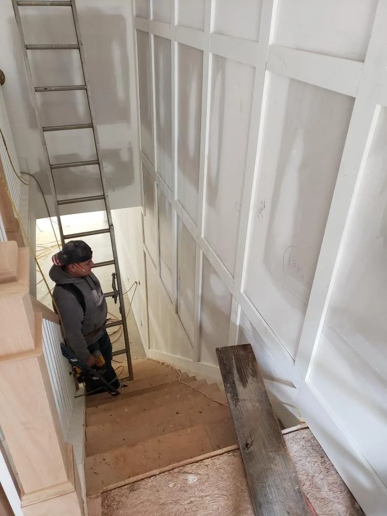 A woman with a black ponytail, wearing glasses, a gray hoodie, and gloves, standing on a staircase inside a house under construction, holding a tool, with an aluminum ladder leaning against the wall.