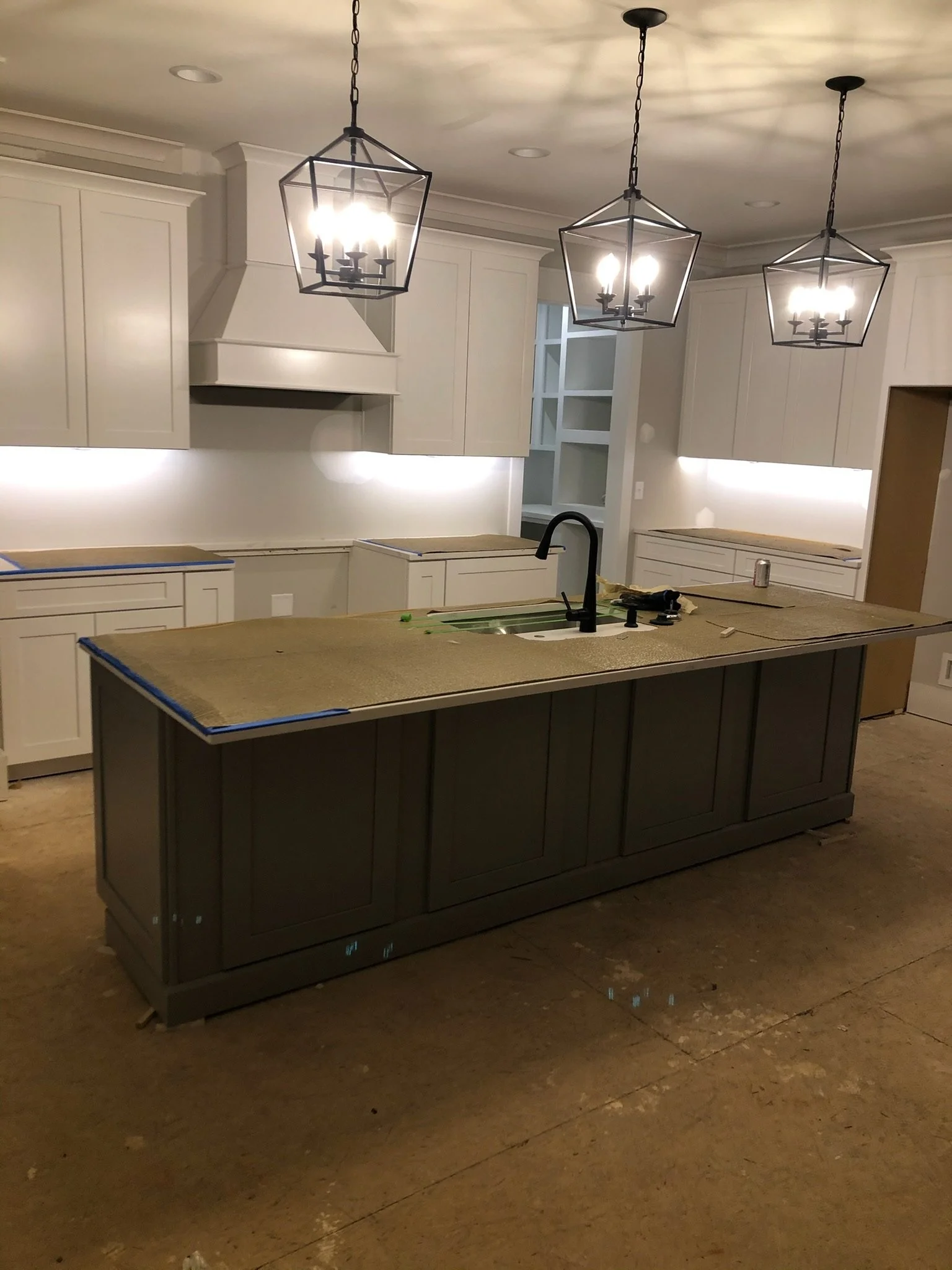 A kitchen with a large island countertop, white cabinets, and three black pendant lights hanging from the ceiling, with some construction materials and tools present.