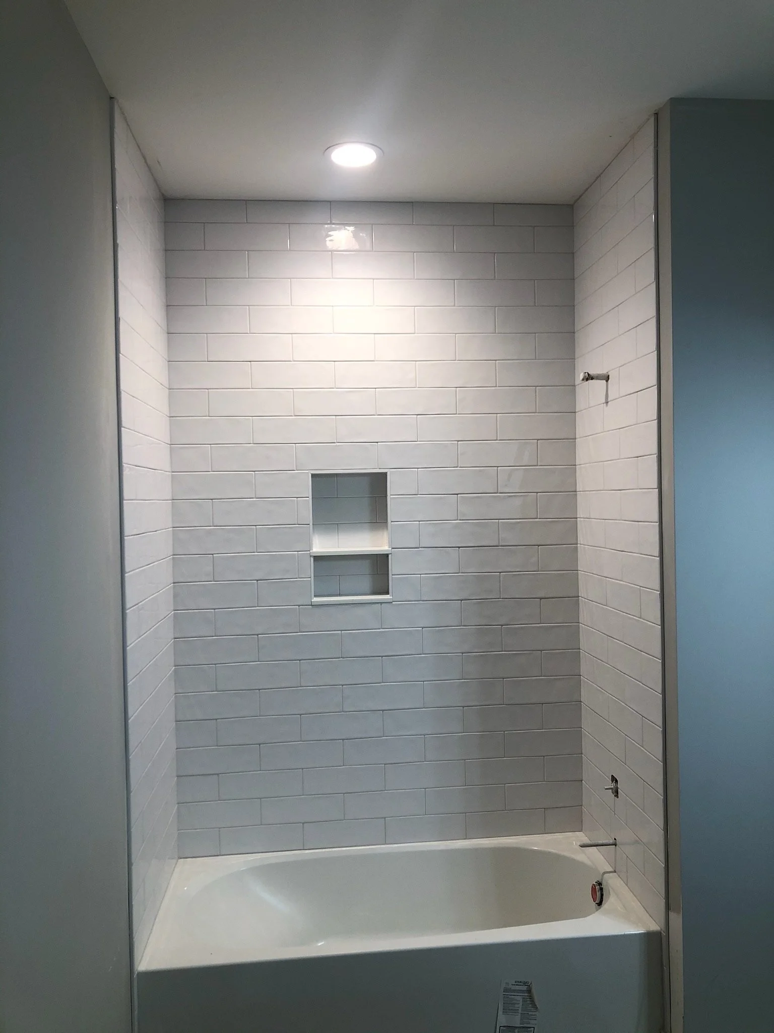A bathroom with white subway tile walls, a built-in niche, a white bathtub, and an overhead light. The plumbing fixtures are not yet installed, with visible pipe connections.
