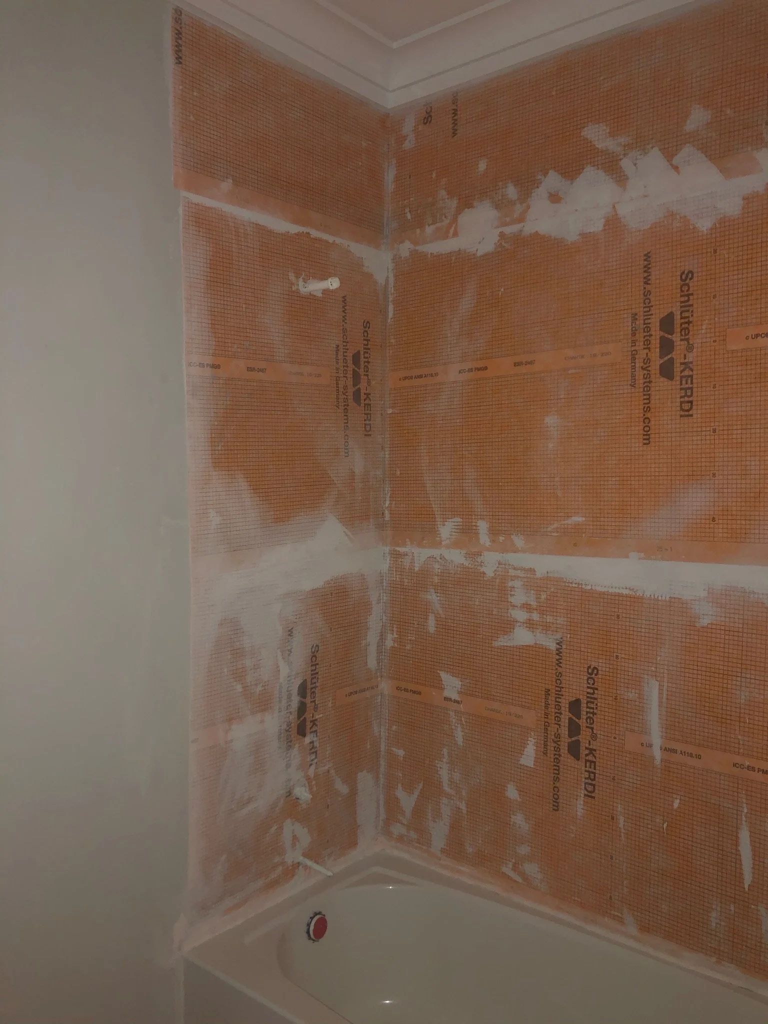 A corner of a bathroom with orange drywall insulation panels and white caulk. The bathtub is white with a red and white faucet handle visible. The walls are not yet finished.