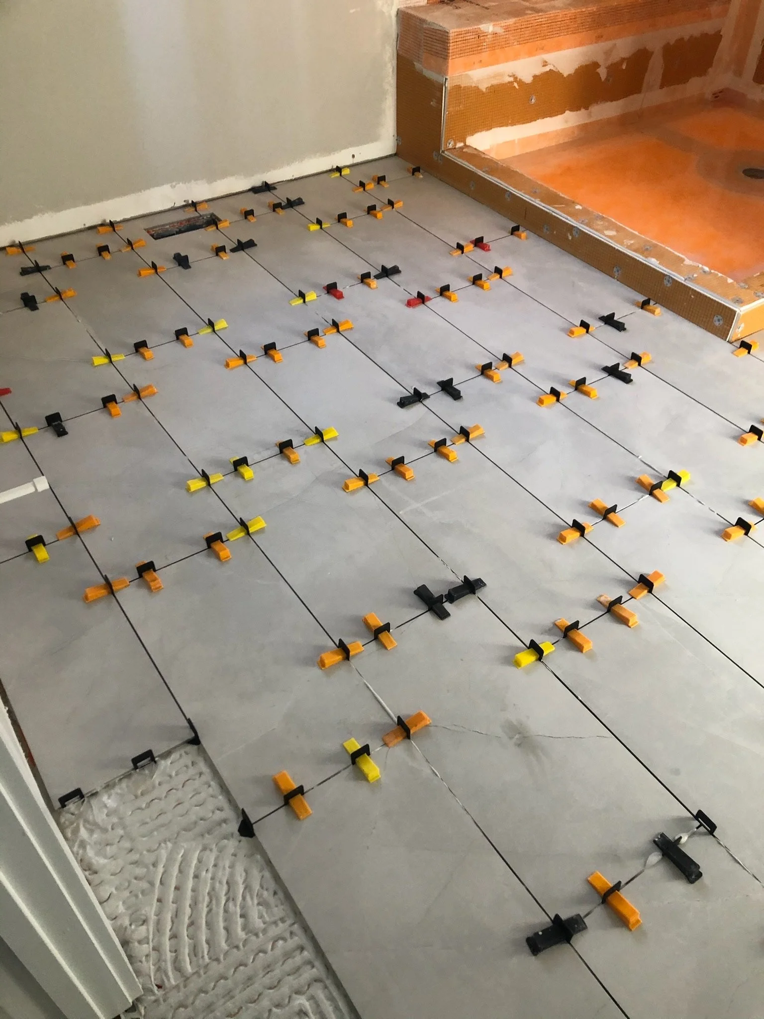 Floor with tile spacers and leveling system during tile installation, near a bathroom shower area with unfinished wall.