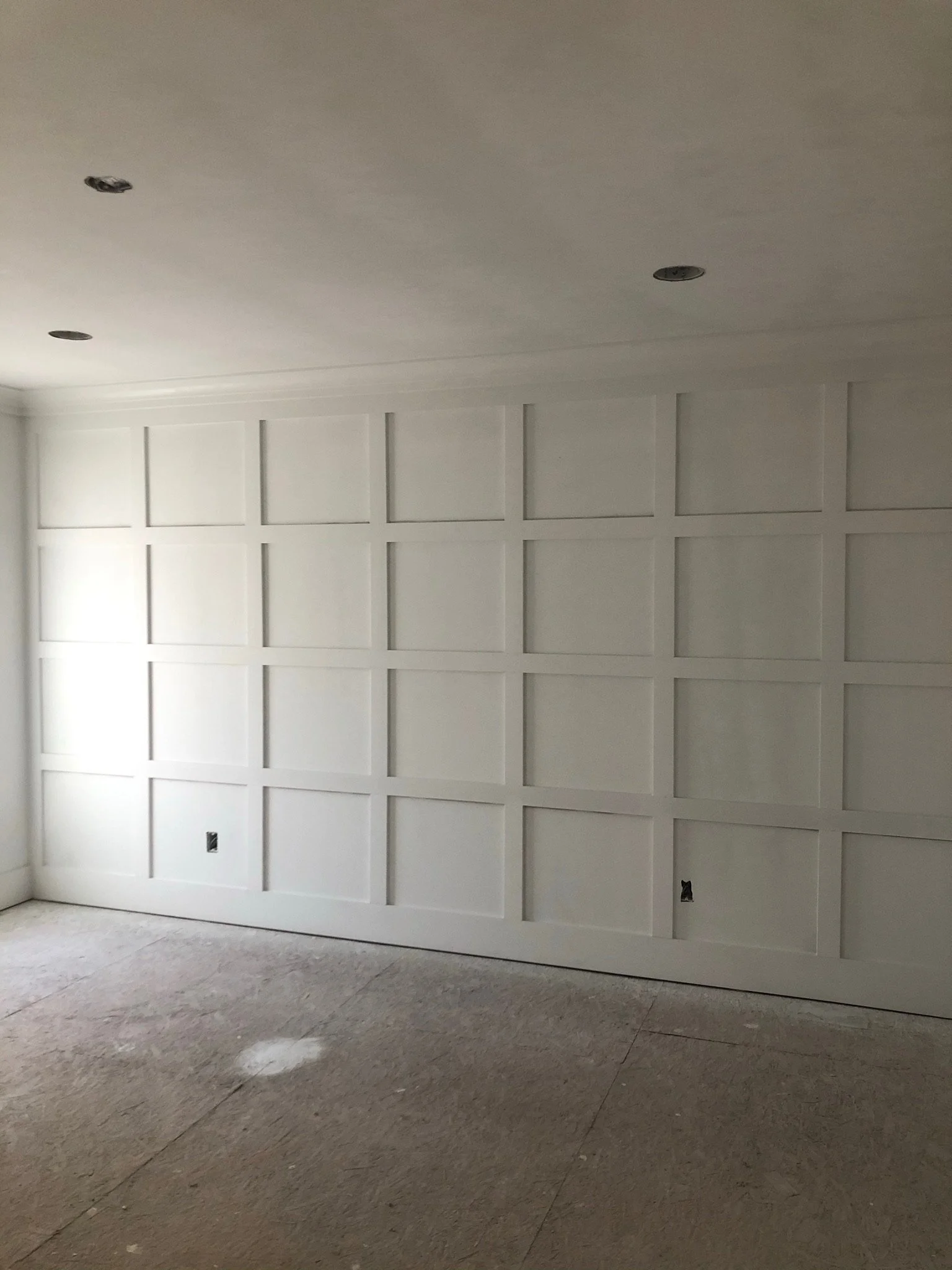 A white wall with decorative rectangular panels and two electrical outlets, with a light-colored carpeted floor in the foreground.
