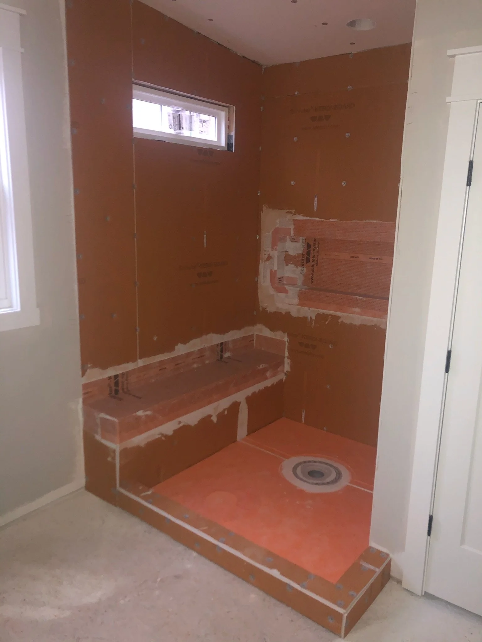 A bathroom shower area with unfinished orange drywall and a small, horizontal window. The floor is unfinished with visible studs and a circular drain.