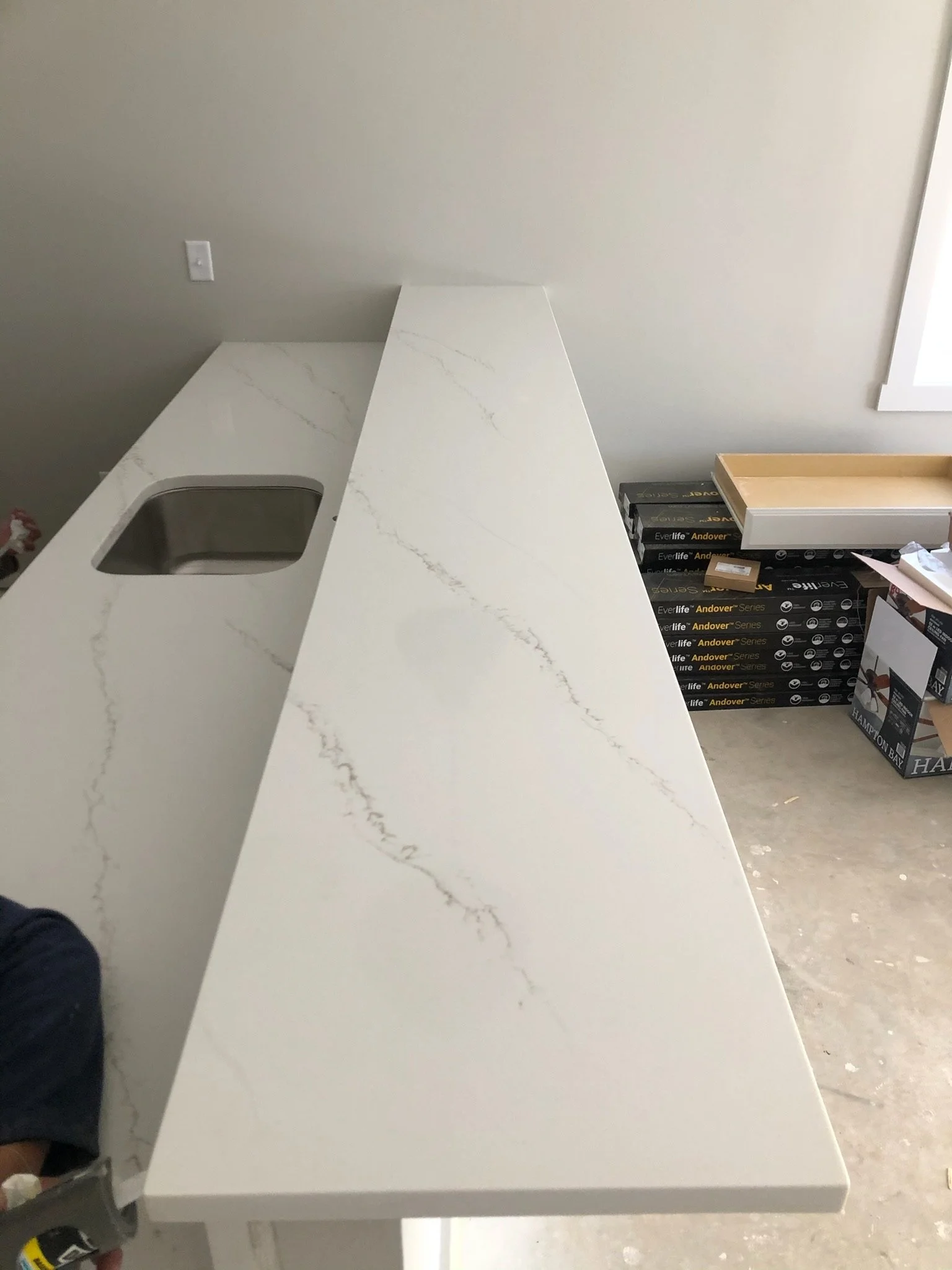 A white marble countertop with gray veining in a room under construction, with boxes and materials stacked against a gray wall and a window on the right.