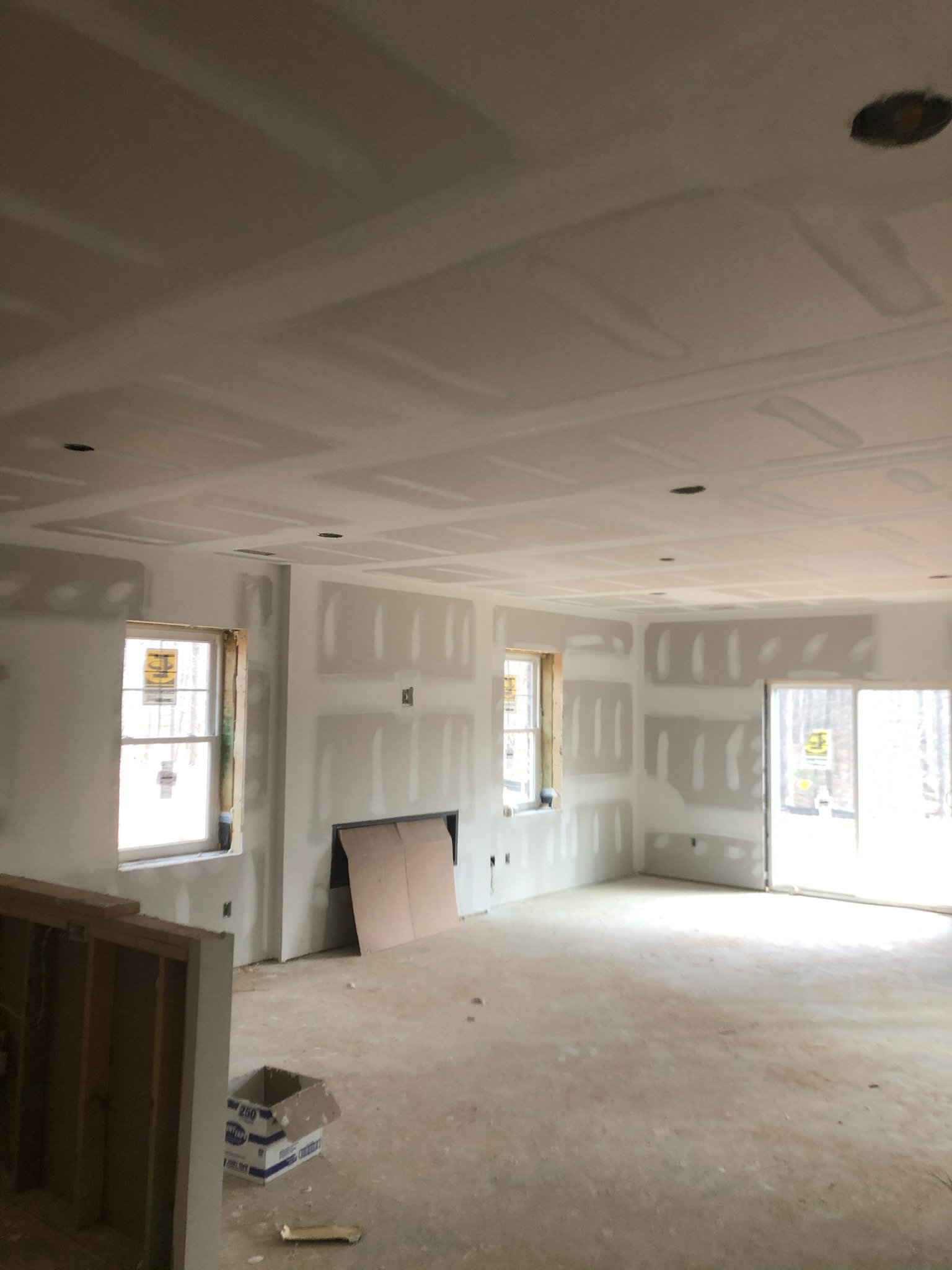 Interior of a building under construction with drywall panels on the walls and ceiling, unfinished floors, and two windows allowing natural light.