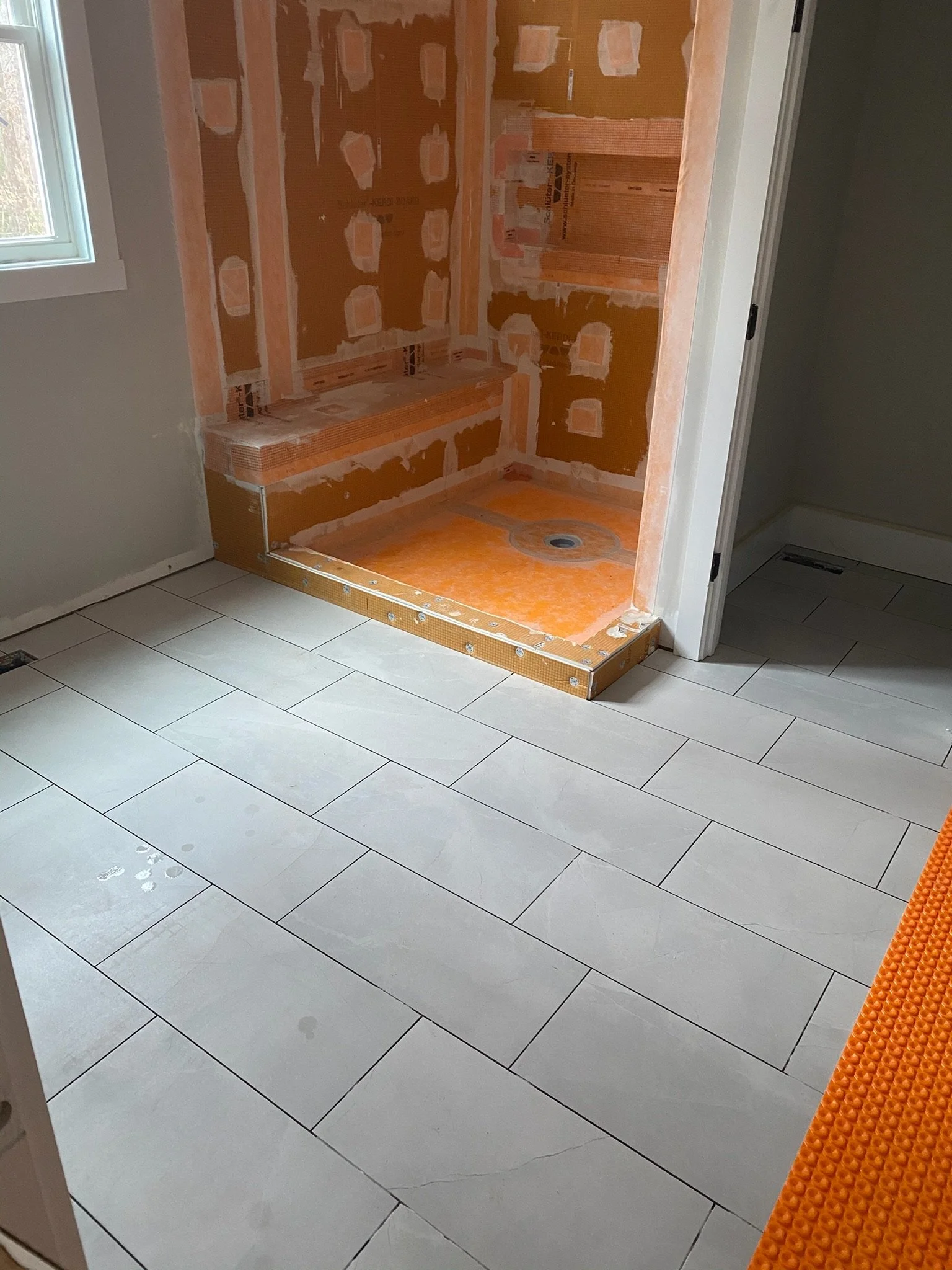 Undergoing bathroom renovation, showing an empty shower stall with orange waterproofing and unfinished walls, adjacent to a white-tiled floor and a window, with a portion of an orange textured mat in the corner.