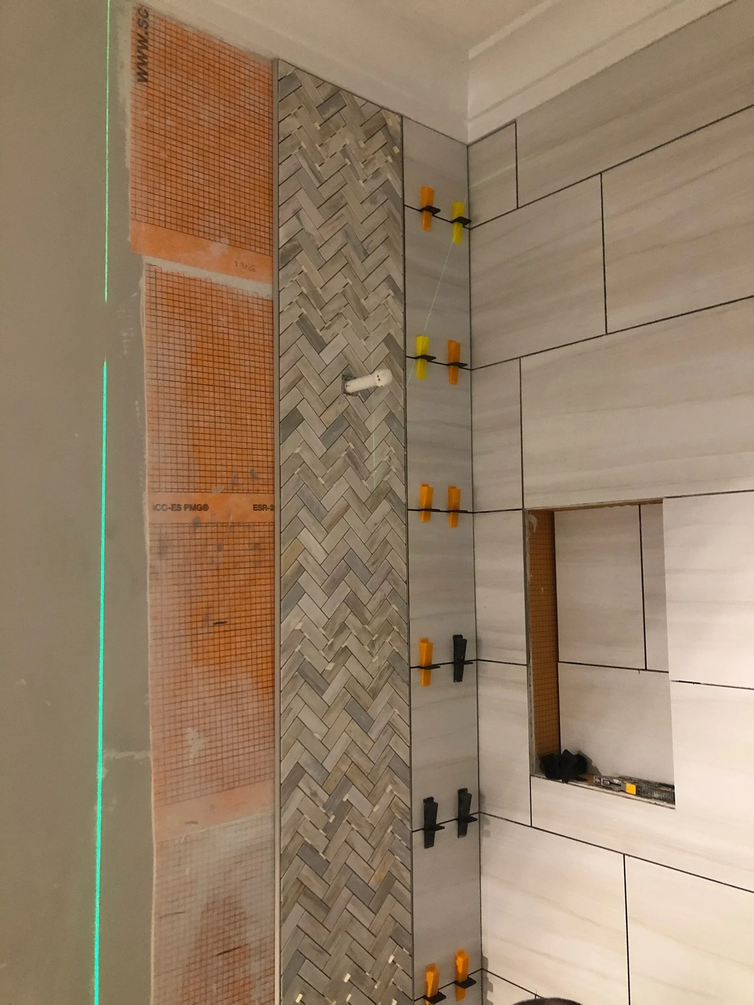 Construction wall with orange waterproof membrane, herringbone wooden pattern, and large light-colored rectangular tiles. Various tools and spacers are placed on the wall. Leveling laser beam projected across the wall.