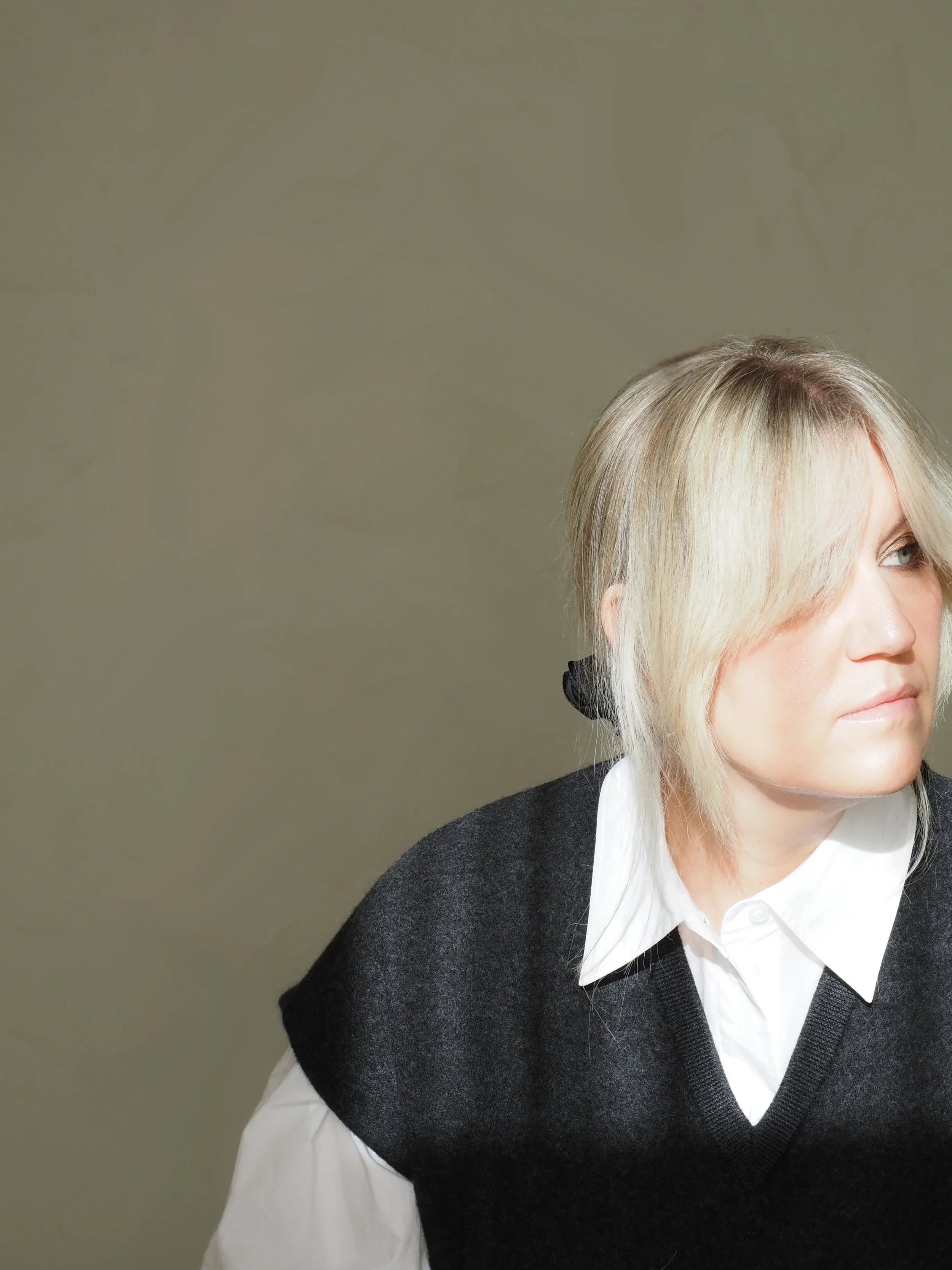 Close-up of a woman with blonde hair and blue eyes wearing a white shirt and black sweater, looking to the right against a neutral background.