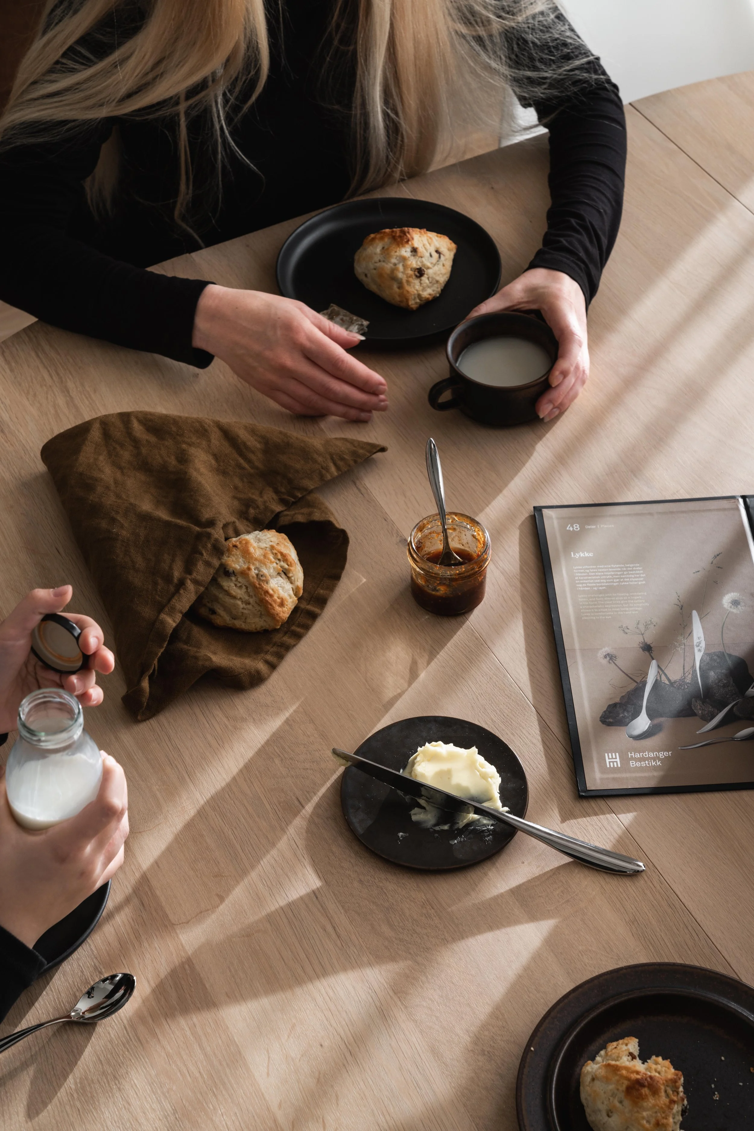 A person with long blonde hair wearing a black long-sleeve shirt sitting at a wooden table with a breakfast meal. The table has a plate with an English scone, a bowl of butter, a jar of jam, a cup of milk, and a glass of milk. There is also a menu or brochure on the table.