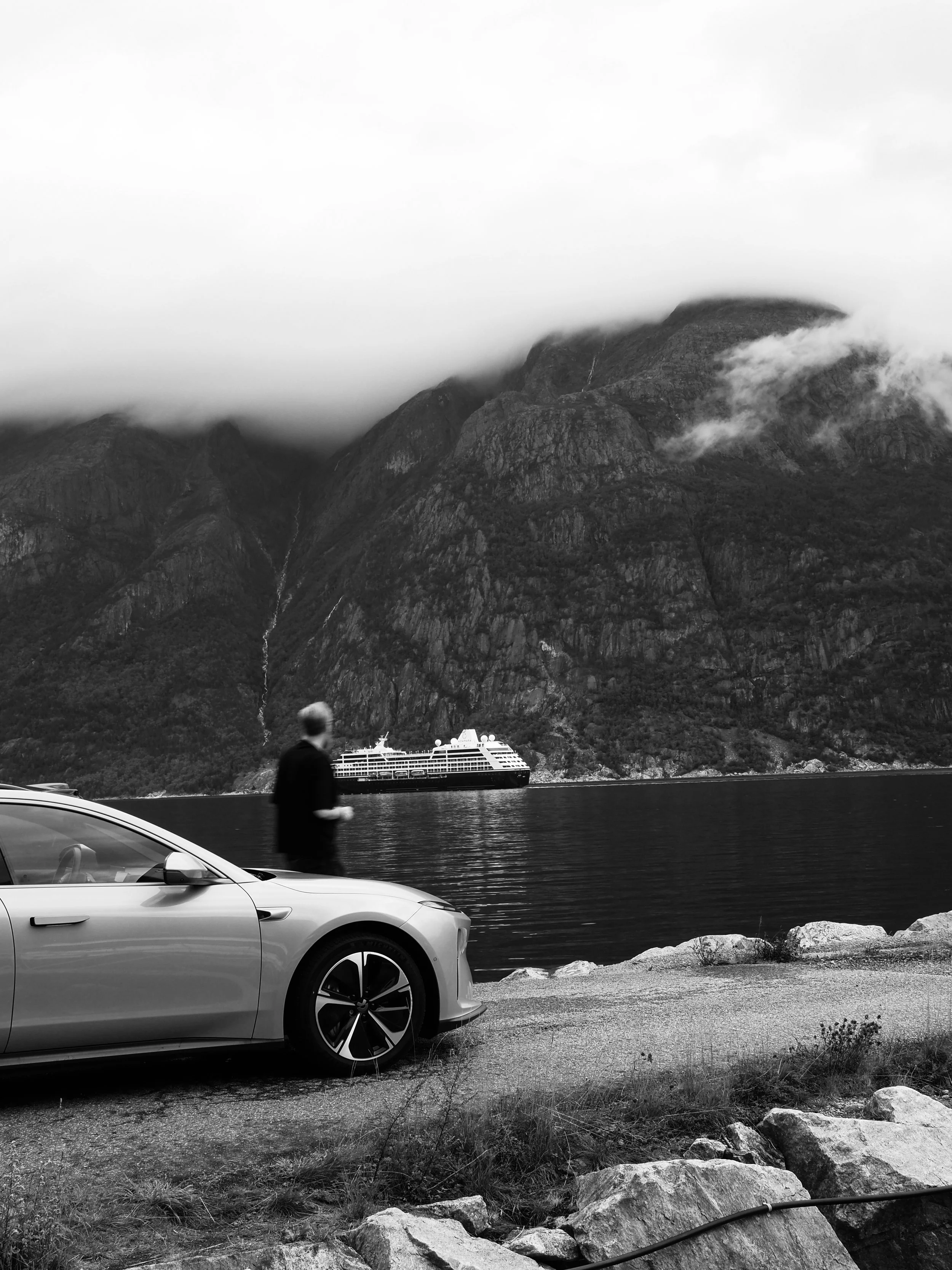 A black and white photo of a mountainous landscape with low-hanging clouds. In the foreground, there are parked cars and a person walking by. A cruise ship is visible on the water at the base of the mountains.