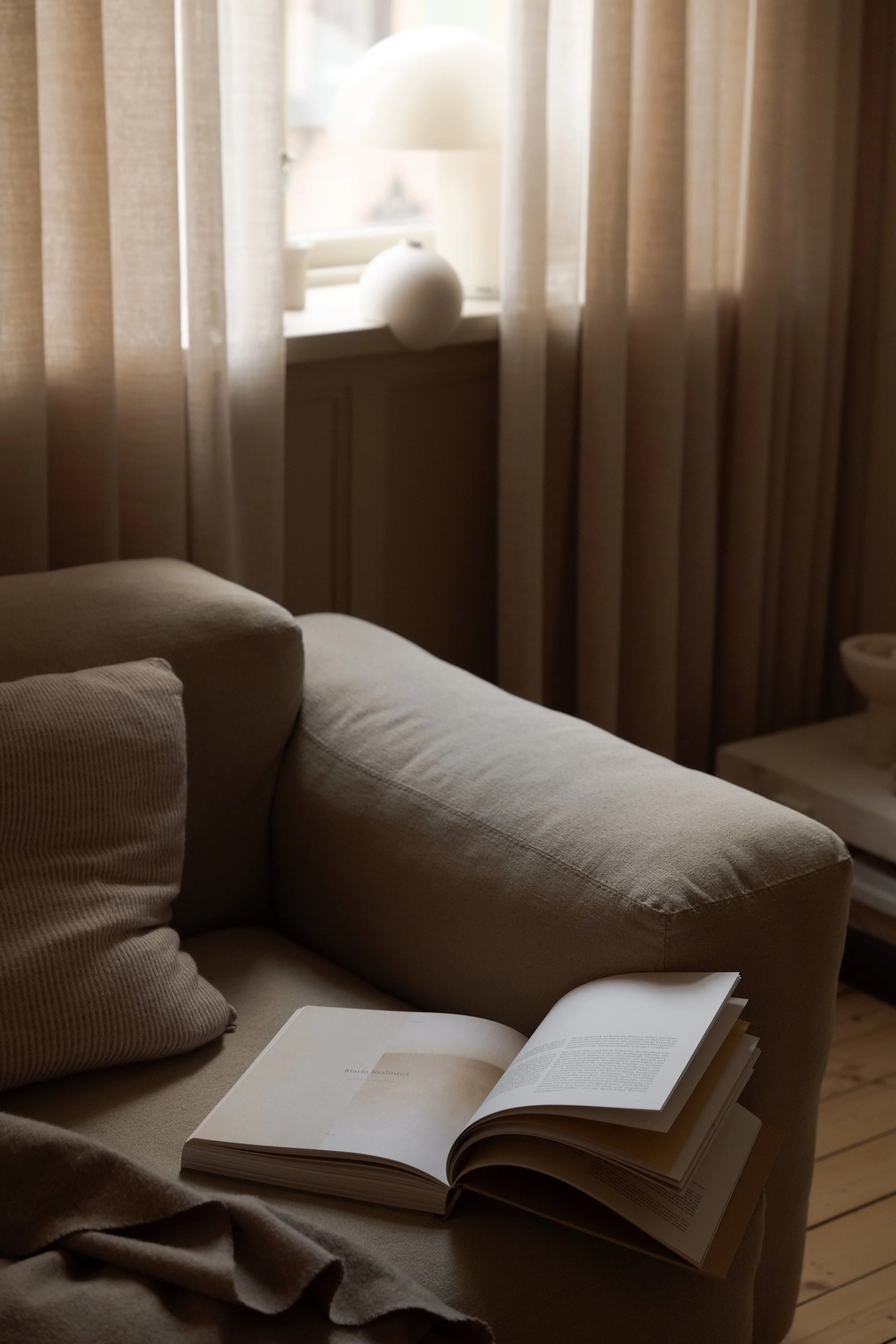 A cozy beige sofa with an open book resting on it, in front of a window with sheer curtains and a decorative white lamp on the windowsill.