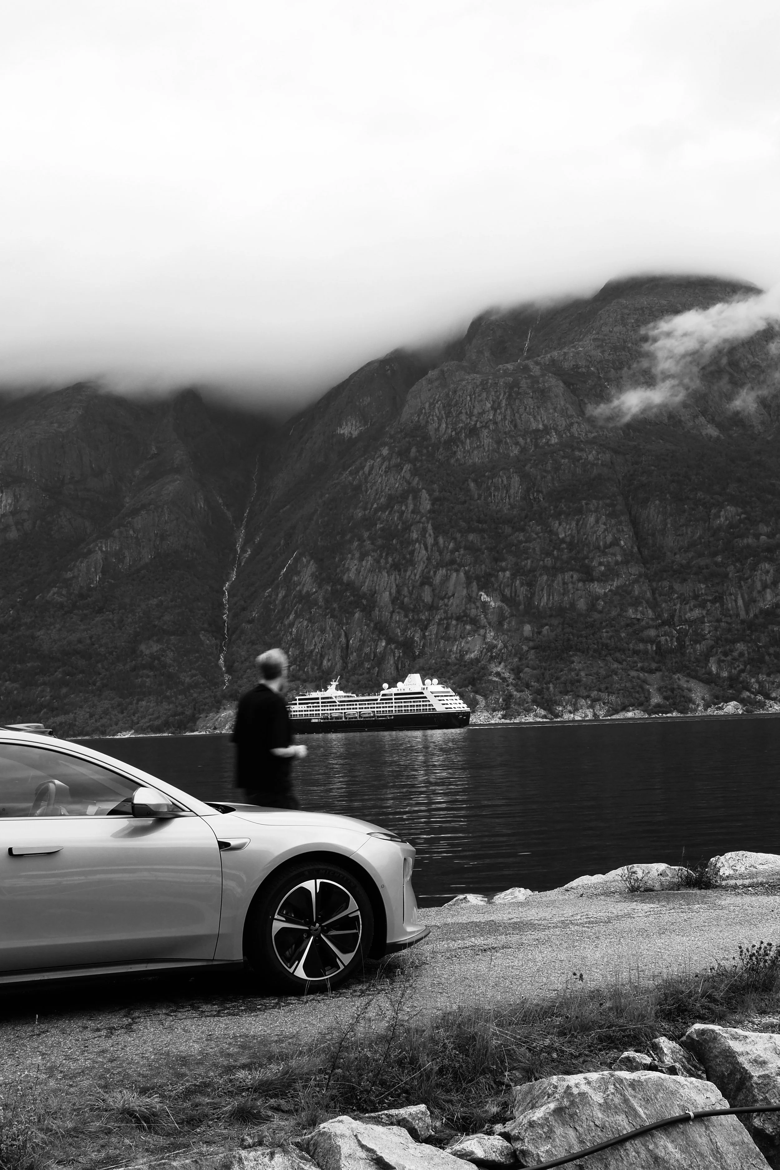 A black-and-white photo of a mountainous landscape with clouds covering the peaks, a cruise ship on the water, a man walking along the shoreline, and part of a car in the foreground.
