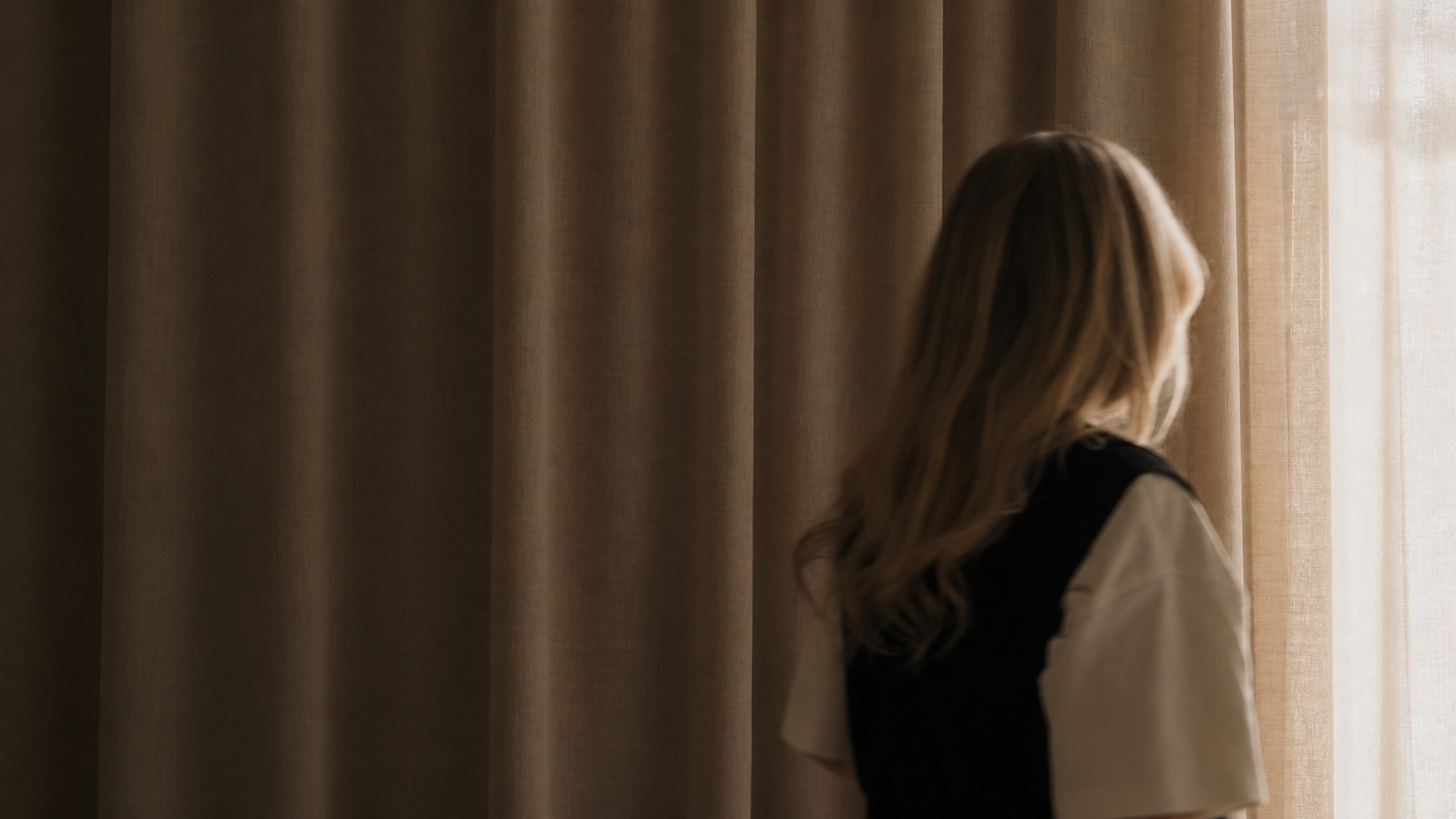 A woman with long, wavy hair looking out a window behind beige curtains, with sunlight illuminating her side.