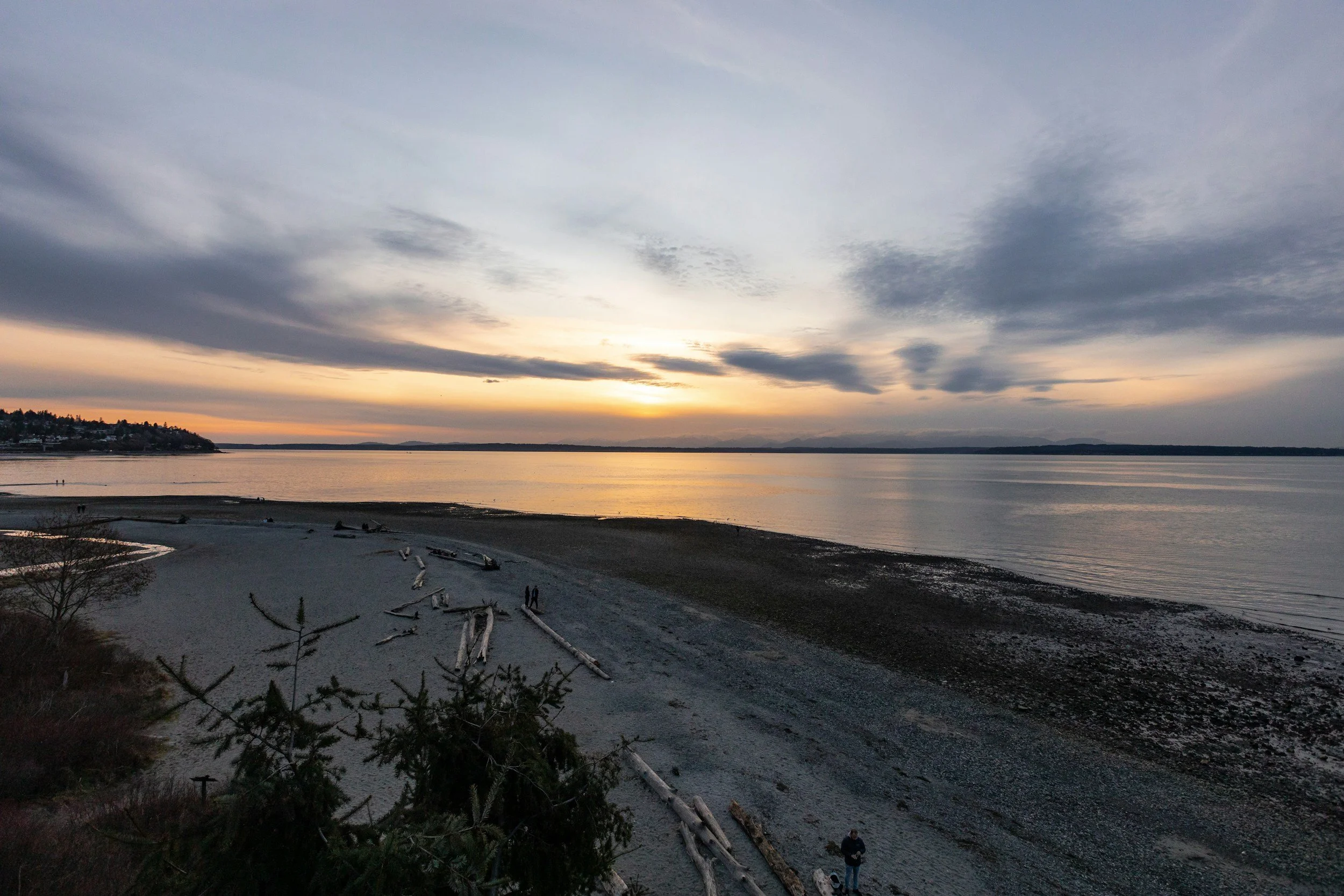 Sunset over a large body of water with a cloudy sky and distant shoreline, beach with driftwood and a few people.